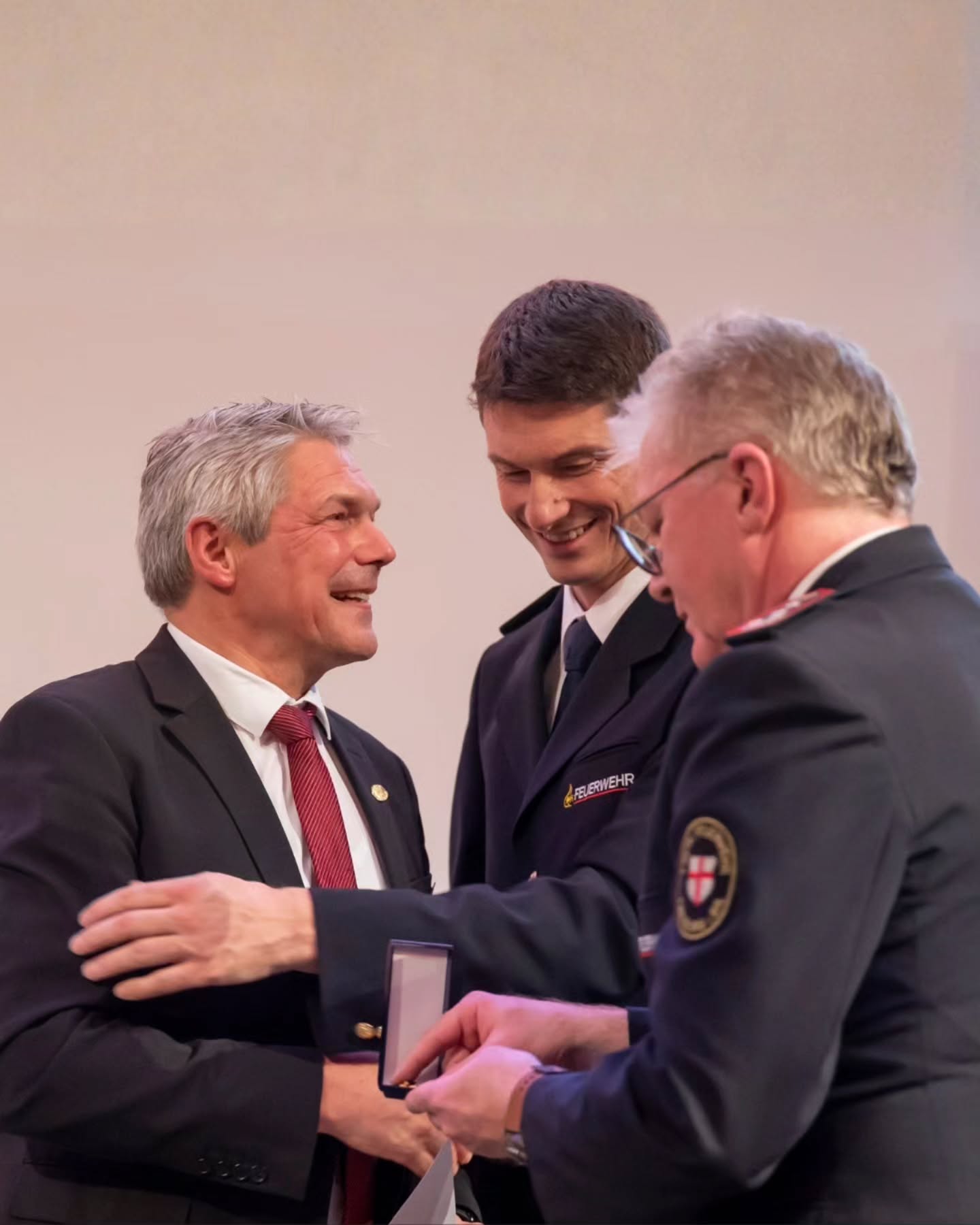 Dieser Alternativtext wurde KI-generiert: Zwei Personen in Anzug und eine Person in Uniform stehen miteinander im Gespräch. Eine Person übergibt eine kleine, dunkle Schachtel.