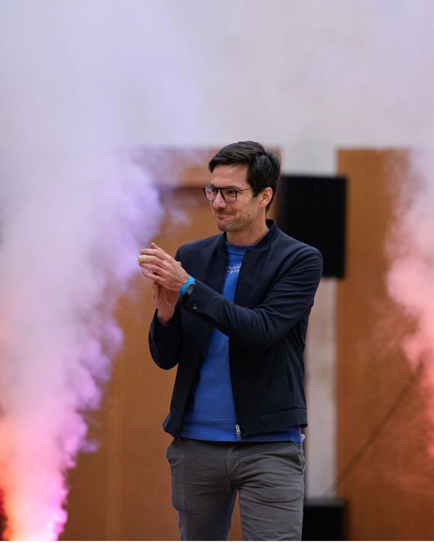 Dieser Alternativtext wurde KI-generiert: Eine Person mit dunklem Haar und Brille steht vor einem Hintergrund mit farbigem Rauch und klatscht in die Hände. Das Bild ist farbig.