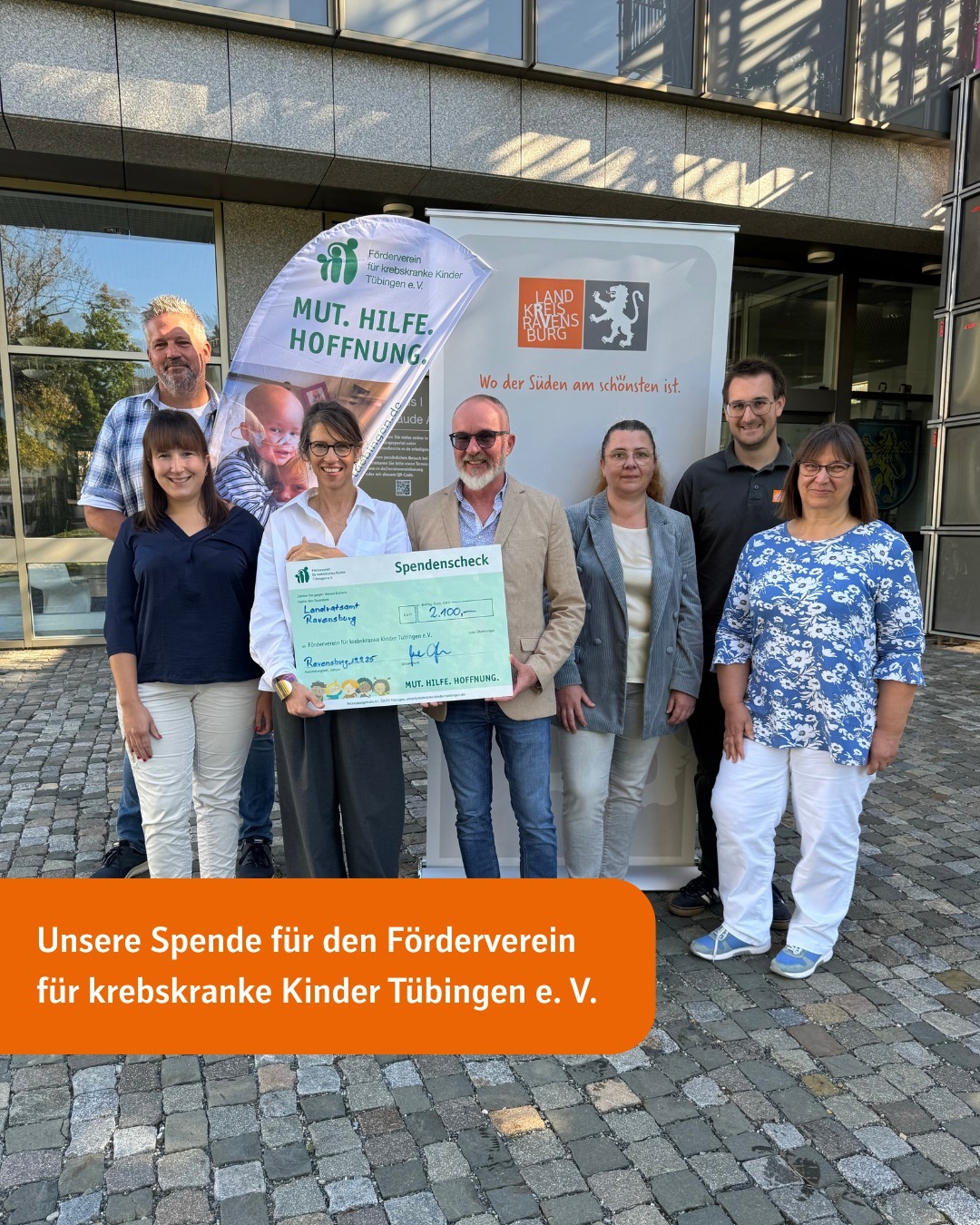 7 Menschen stehen nebeneinander vor dem Landratsamtsgebäude in Ravensburg. Zwei davon halten einen großen Spendenscheck in der Hand. Text im Bild: Unsere Spende für den Förderverein für krebskranke Kinder Tübingen e. V.