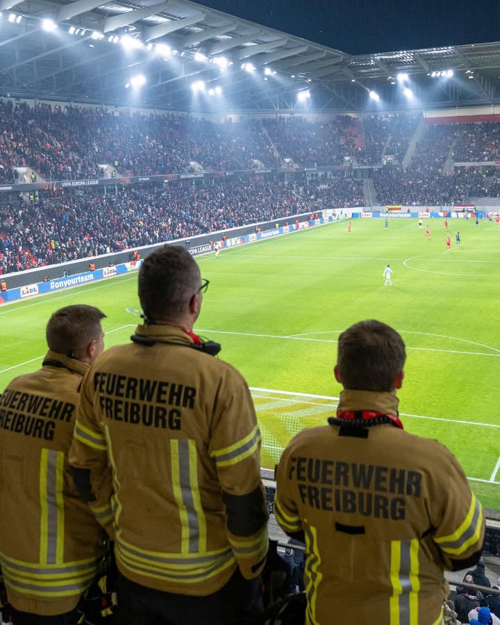 Dieser Alternativtext wurde KI-generiert: Ein Fußballstadion bei Nacht, beleuchtet von starken Scheinwerfern. Im Vordergrund stehen Personen in gelben Uniformen mit der Aufschrift 'FEUERWEHR FREIBURG', die zum Spielfeld blicken.