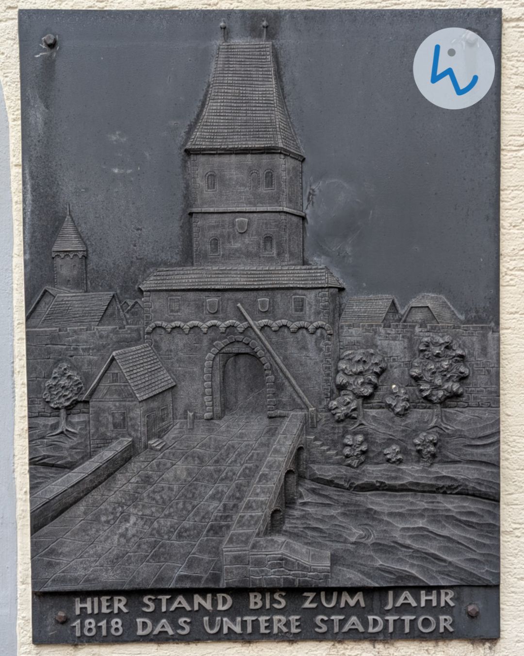 Ein dunkles Metallrelief an einer Wand zeigt ein historisches Stadttor mit Turm, das über eine Brücke führt, flankiert von weiteren Gebäuden und Bäumen. Am unteren Rand der Tafel steht der Text: „HIER STAND BIS ZUM JAHR 1818 DAS UNTERE STADTTOR“. (KI-Alternativtext)