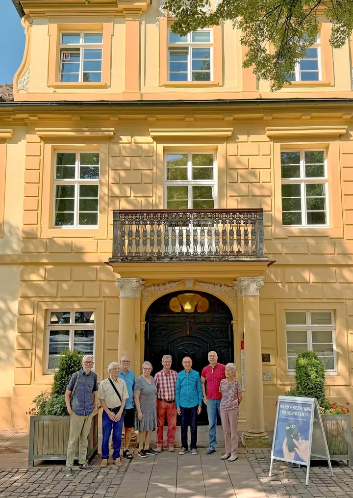 Eine Gruppe von acht Personen steht vor dem Eingang eines gelben Gebäudes mit Balkon. Rechts im Vordergrund steht ein Ständer mit einem Schild, auf dem "HANDY-BERATUNG FÜR SENIOREN" zu lesen ist. (KI-Alternativtext)