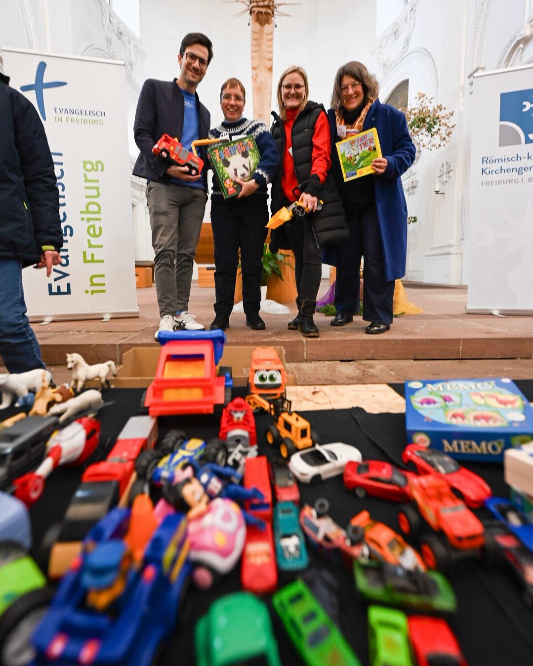 Dieser Alternativtext wurde KI-generiert: Mehrere Personen stehen vor einer Stufe und halten Spielzeug in den Händen. Im Vordergrund befindet sich eine Sammlung von Spielzeugautos und -figuren.