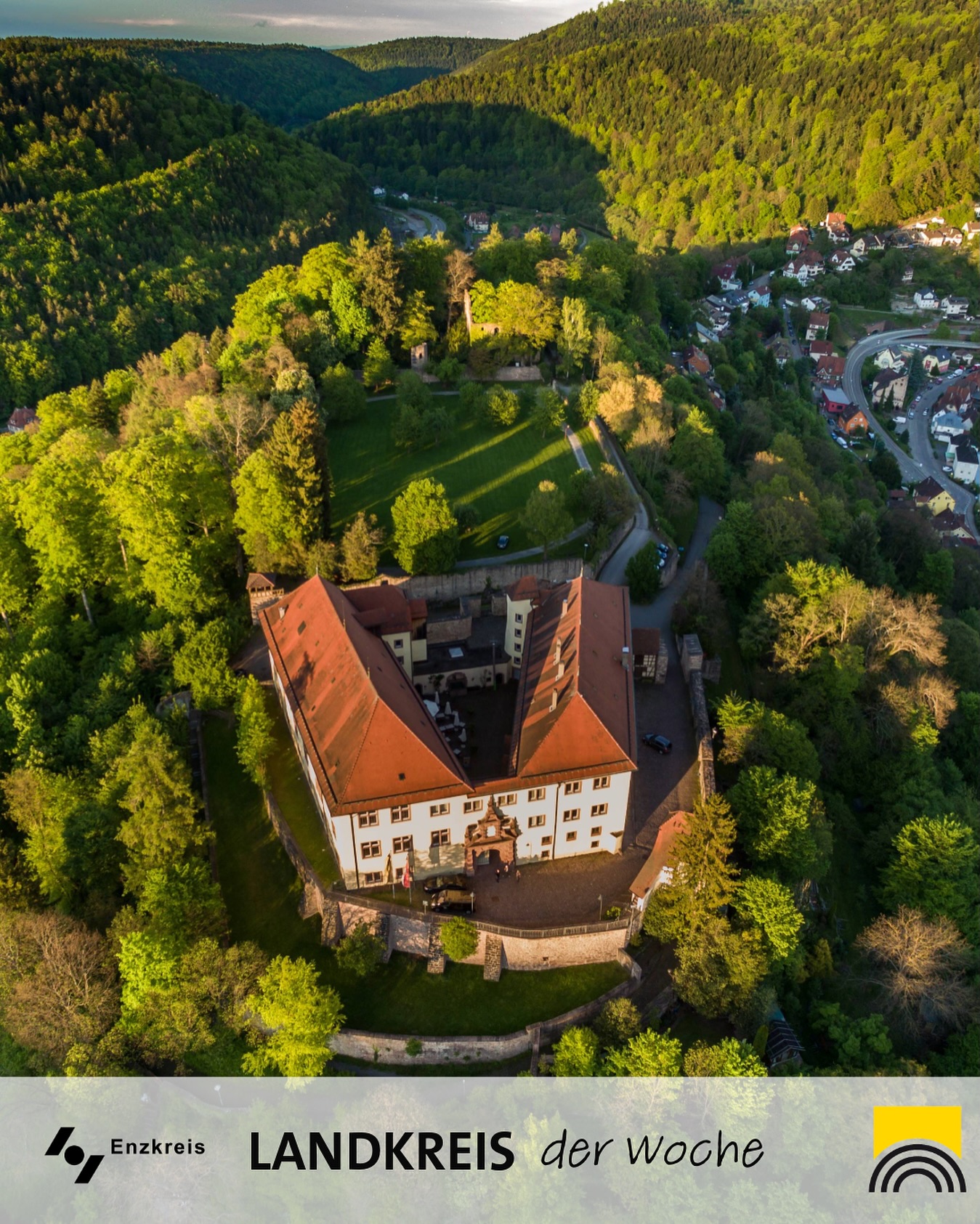 Dieser Alternativtext wurde KI-generiert: Eine Vogelperspektive zeigt ein Gebäude mit roten Ziegeldächern, umgeben von üppigem Grün und einer Stadt. Das Bild ist farbig und wirkt durch das Licht warm.