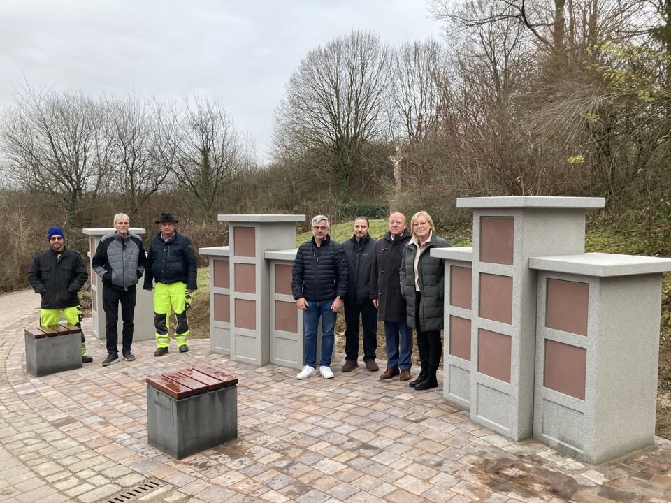 Dieser Alternativtext wurde KI-generiert: Eine Gruppe von Menschen steht vor einer Steinmauer und -konstruktionen in einer Außenanlage. Im Hintergrund sind Bäume und Büsche zu sehen.