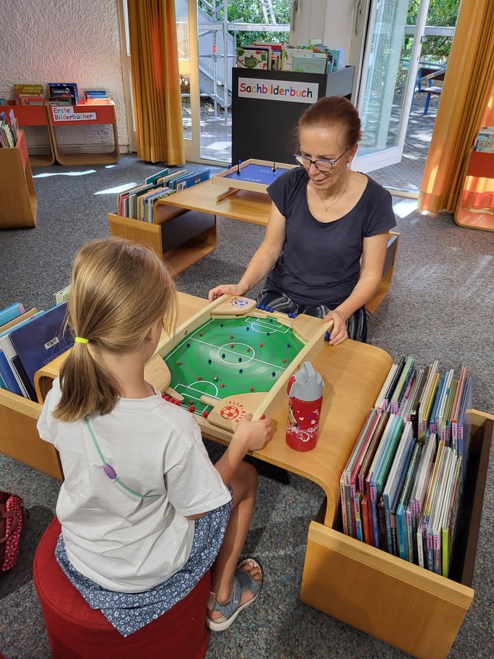 Zwei Personen spielen an einem Holztisch Tischfußball in einem Raum mit Bücherregalen. Im Hintergrund stehen Regale mit den Beschriftungen "Sachbilderbuch" und "Große Bilderbücher". Eine rote Trinkflasche steht auf dem Tisch. (KI-Alternativtext)