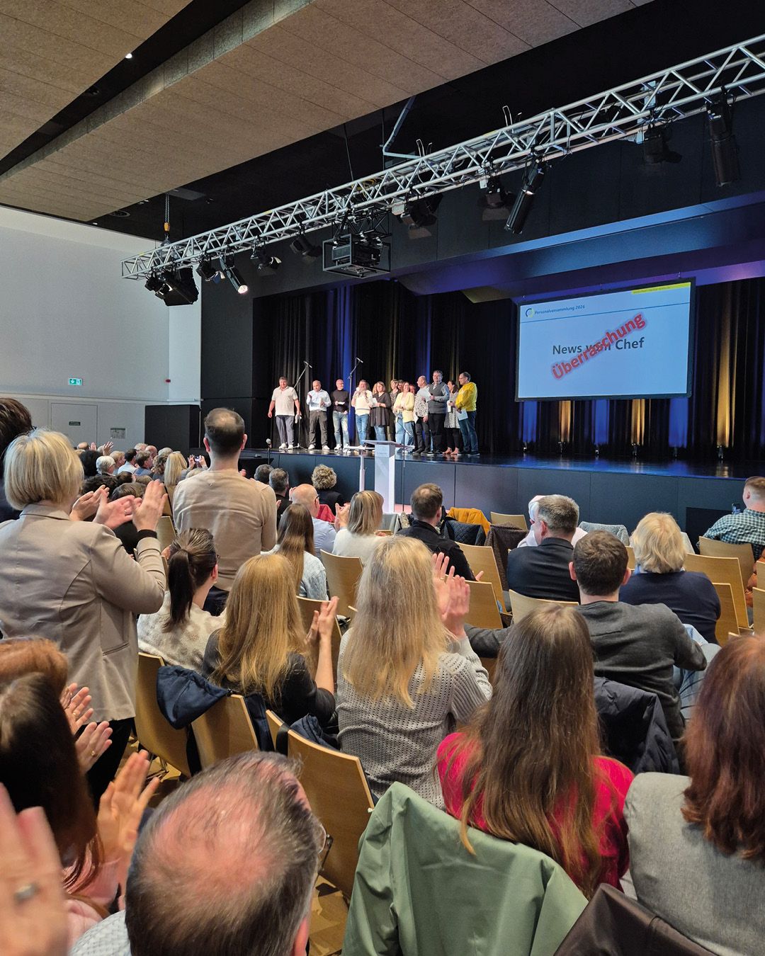 Dieser Alternativtext wurde KI-generiert: Eine Gruppe von Personen steht auf einer Bühne vor einem Publikum. Im Hintergrund befindet sich eine Bühne mit technischer Ausstattung und eine Präsentation auf einer Leinwand.