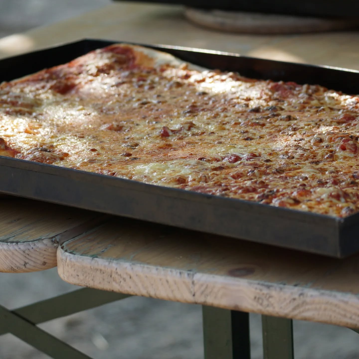 Dieser Alternativtext wurde KI-generiert: Ein großes, rechteckiges Blech mit einer Käse-Pizza steht auf einem Holztisch im Freien. Die Pizza ist mit verschiedenen Belägen belegt.