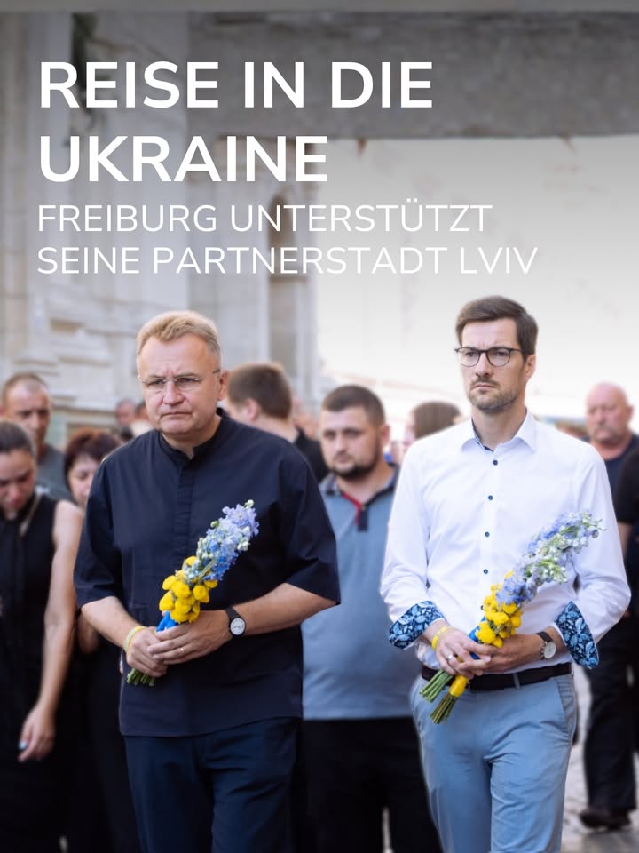 Zwei Personen stehen im Vordergrund und halten Sträuße mit gelben und blauen Blumen, während im Hintergrund weitere Personen zu sehen sind. Der Text "REISE IN DIE UKRAINE" und "FREIBURG UNTERSTÜTZT SEINE PARTNERSTADT LVIV" ist über dem Bild platziert. (KI-Alternativtext)