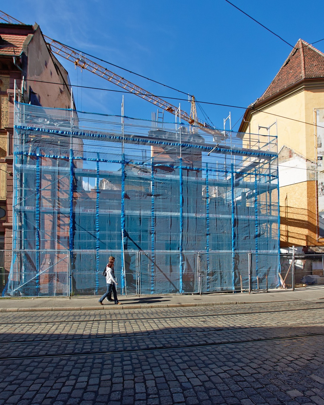 Dieser Alternativtext wurde KI-generiert: Ein Gebäude ist von Gerüsten umgeben, die mit blauen Planen verkleidet sind. Eine Person geht auf einem gepflasterten Platz an dem Bau vorbei.