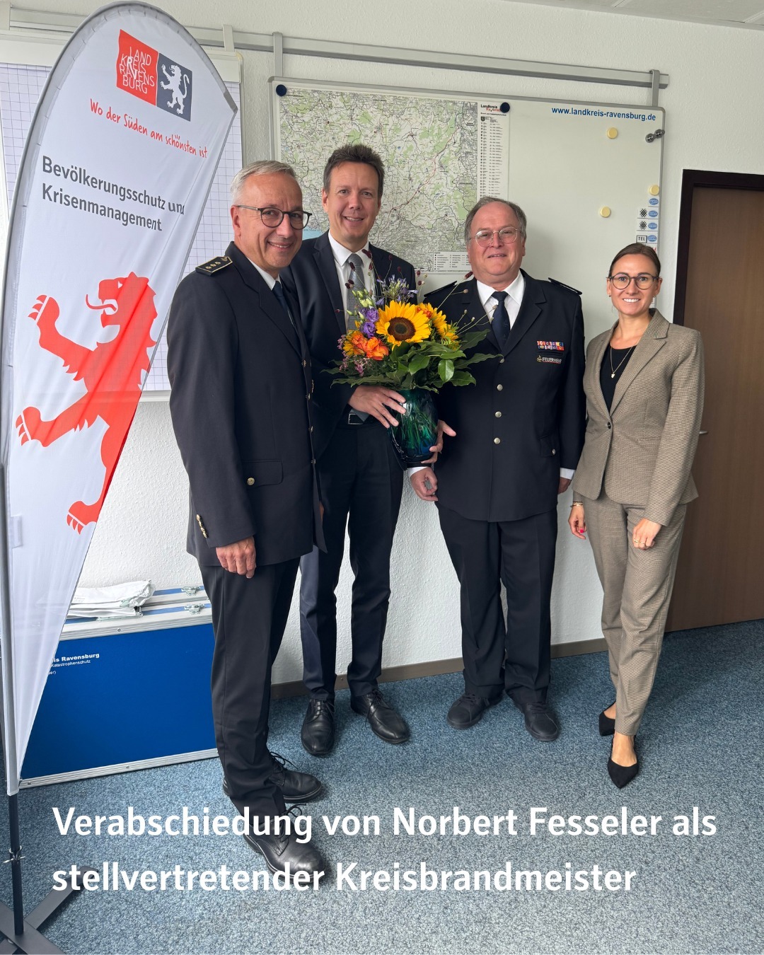 Vier Personen stehen vor einer Wand mit einer Karte. Eine Person hält einen Blumenstrauß.