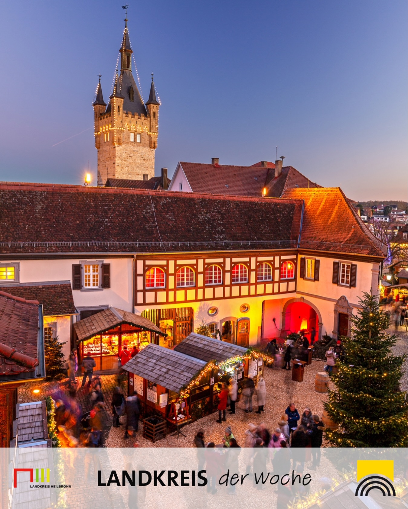 Dieser Alternativtext wurde KI-generiert: Ein farbiges Bild zeigt eine Stadtansicht mit historischen Gebäuden und einem beleuchteten Turm. Im Vordergrund befindet sich ein Weihnachtsmarkt mit Ständen und Besuchern.