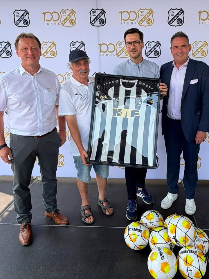 Vier Personen stehen vor einem Hintergrundbanner mit dem Logo des SV Mühlburg und der Aufschrift „100 Jahre“. Zwei Personen in der Mitte halten ein gerahmtes, schwarz-weiß gestreiftes Trikot mit der Aufschrift „KIF“ sowie den Jahren 1926 und 2026. Auf dem Boden im Vordergrund liegen mehrere Fußball. (KI-Alternativtext)