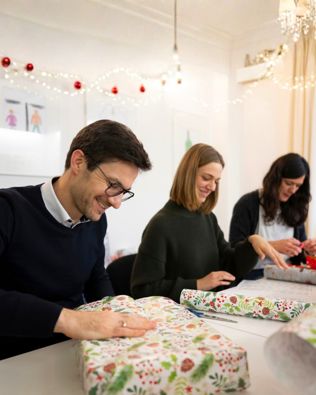 Dieser Alternativtext wurde KI-generiert: Mehrere Personen sitzen an einem Tisch und verpacken Geschenke in festlicher Atmosphäre. Im Hintergrund sind Weihnachtsdekorationen und Lichter zu sehen.