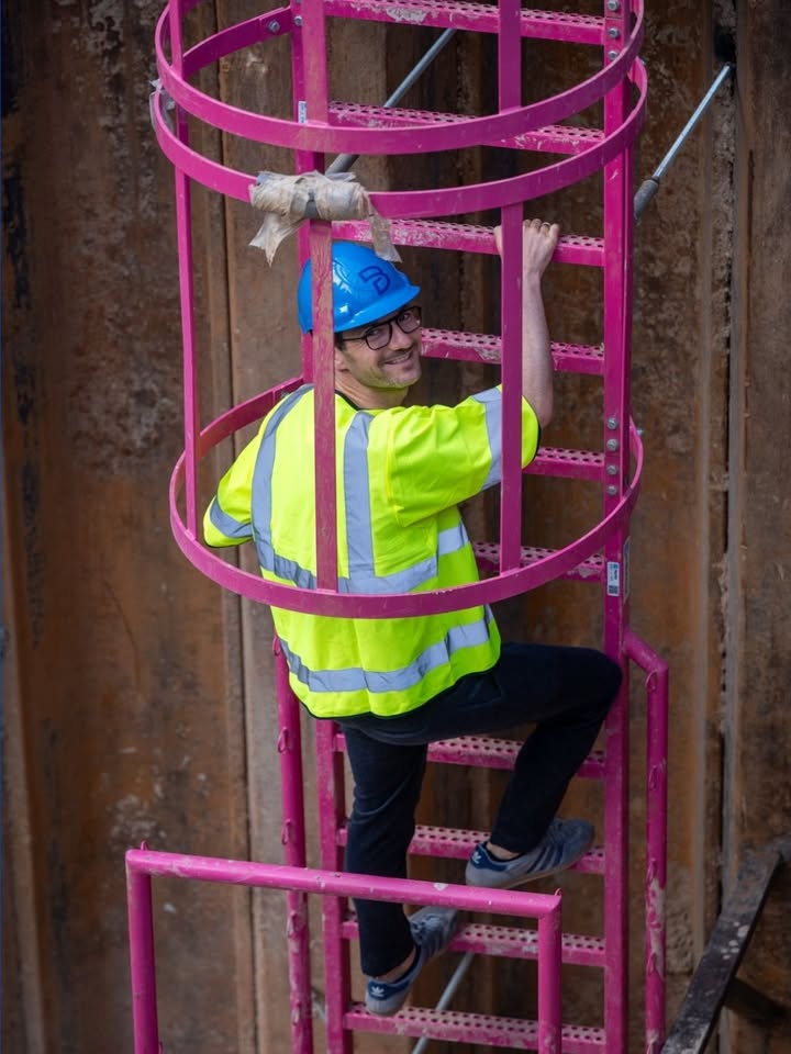 Eine Person in neongelber Warnweste und blauem Schutzhelm klettert eine rosa Leiter innerhalb eines Schutzgitters hinauf. Die Person blickt lächelnd zurück. Im Hintergrund ist eine Holzschalung zu sehen. (KI-Alternativtext)