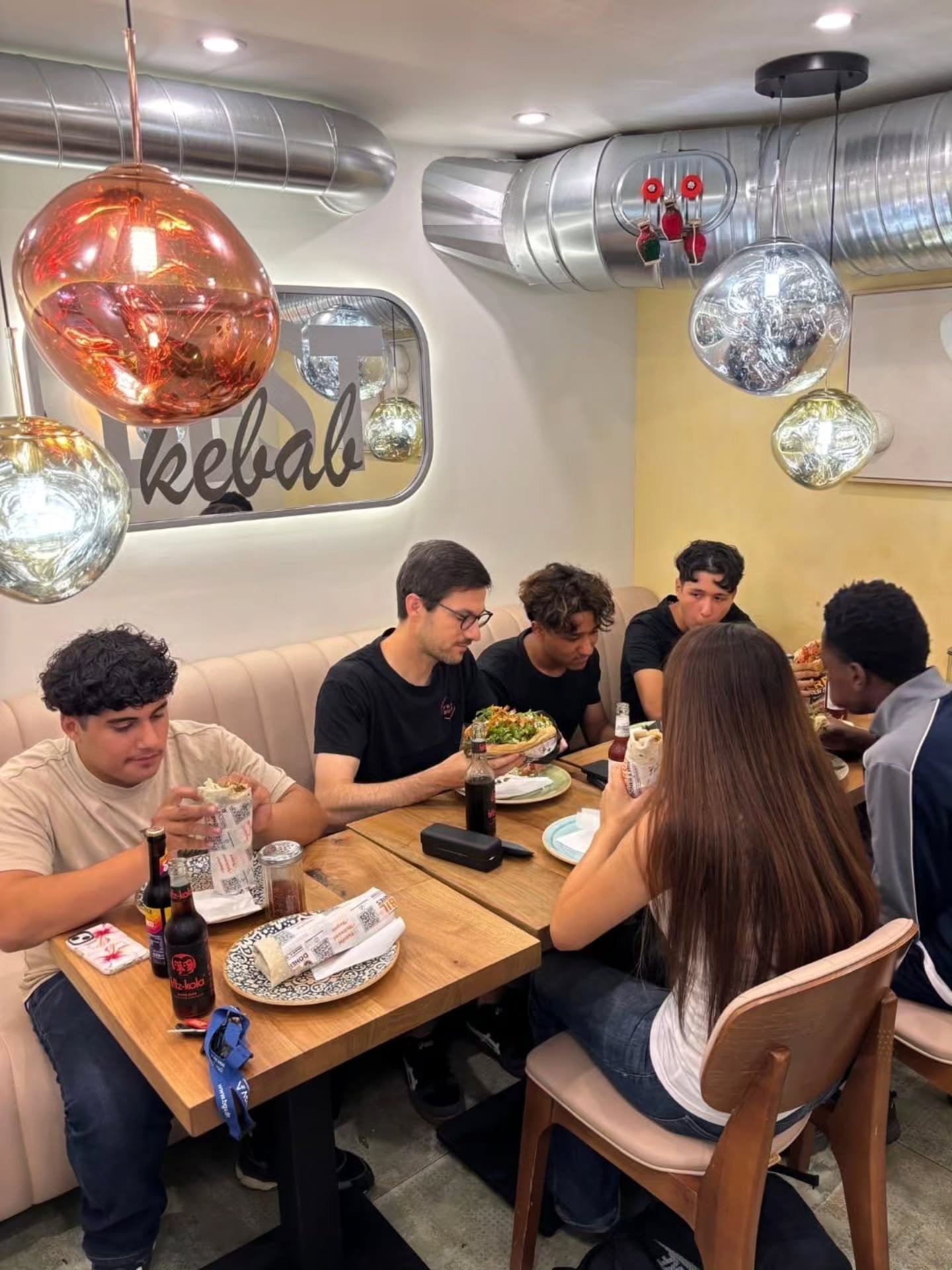 Eine Gruppe von Personen isst an Holztischen in einem Restaurant, wobei Wraps und Salate auf den Tellern liegen. Flaschen mit dunkler Limonade stehen auf dem Tisch. Im Hintergrund ist ein Schild mit der Aufschrift "kebab" sowie silberne Lüftungskanäle und kugelförmige Pendelleuchten an der Decke zu erkennen. (KI-Alternativtext)