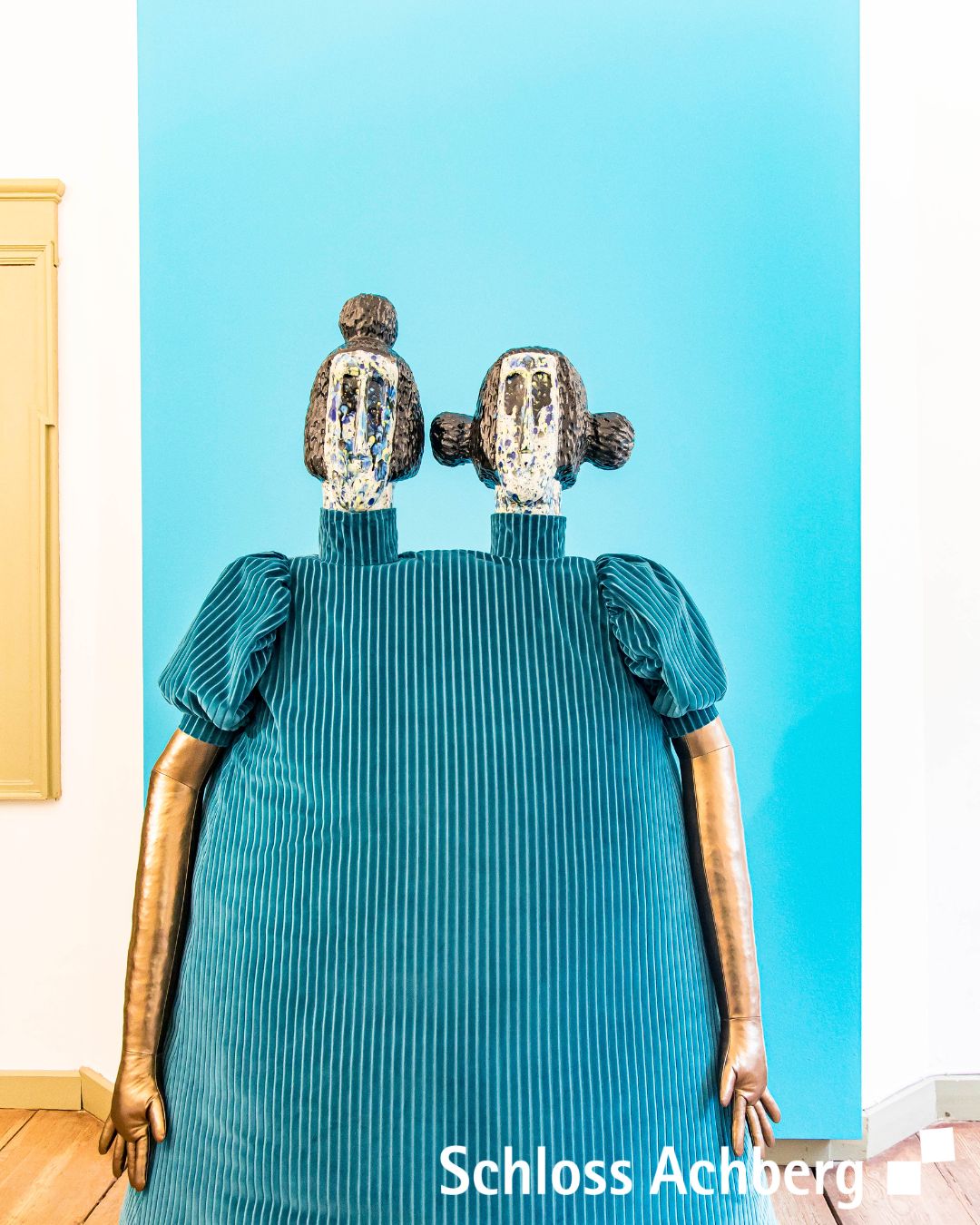 Eine farbenfrohe Skulptur, die an zwei Figuren in Kleidung erinnert, steht vor einer blauen Wand. Die Skulptur hat einen stilisierten Kopf und Arme.