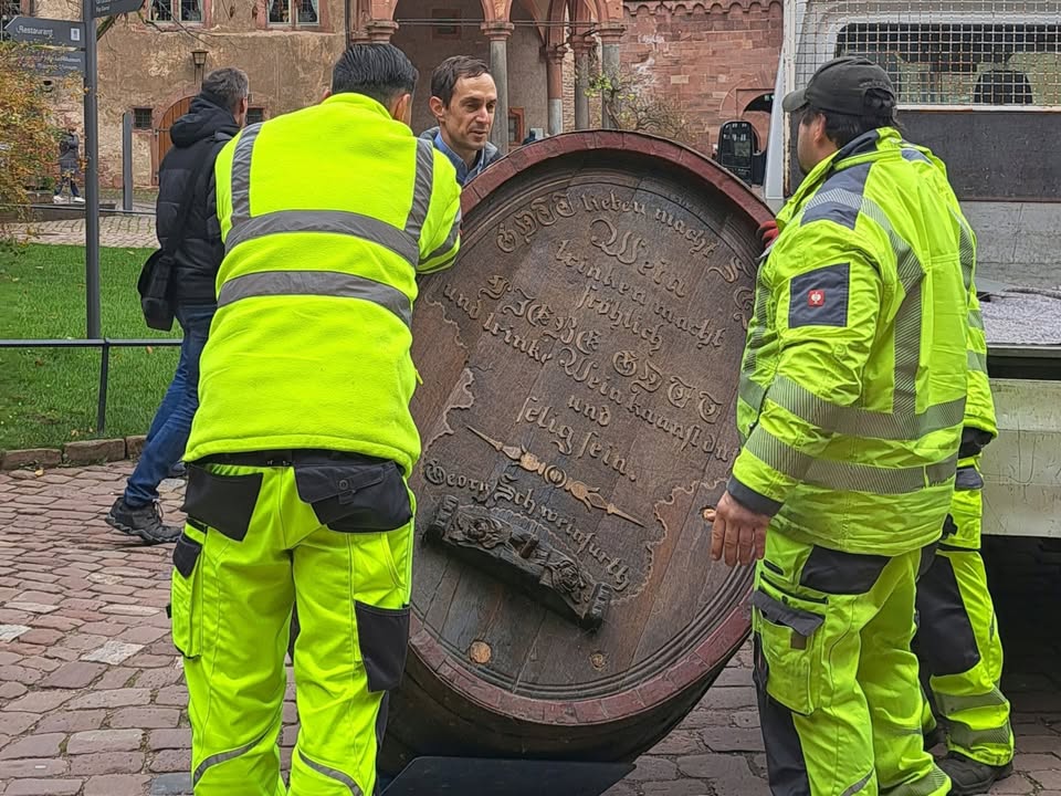 Dieser Alternativtext wurde KI-generiert: Mehrere Personen in heller Kleidung bewegen ein großes, historisch aussehendes Holzfass mit Beschriftungen auf einem gepflasterten Platz. Im Hintergrund ist ein Gebäude mit verzierten Fassadenelementen zu sehen.