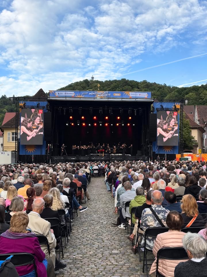 Große Menschenmenge sitzt auf Stühlen vor einer Bühne mit Orchester und großen Bildschirmen, auf denen Musikerinnen zu sehen sind. Im Hintergrund grüner Hügel und blauer Himmel mit Wolken. Auf der Bühne steht „Münsterplatz Konzerte 2024“. (KI-Alternativtext)