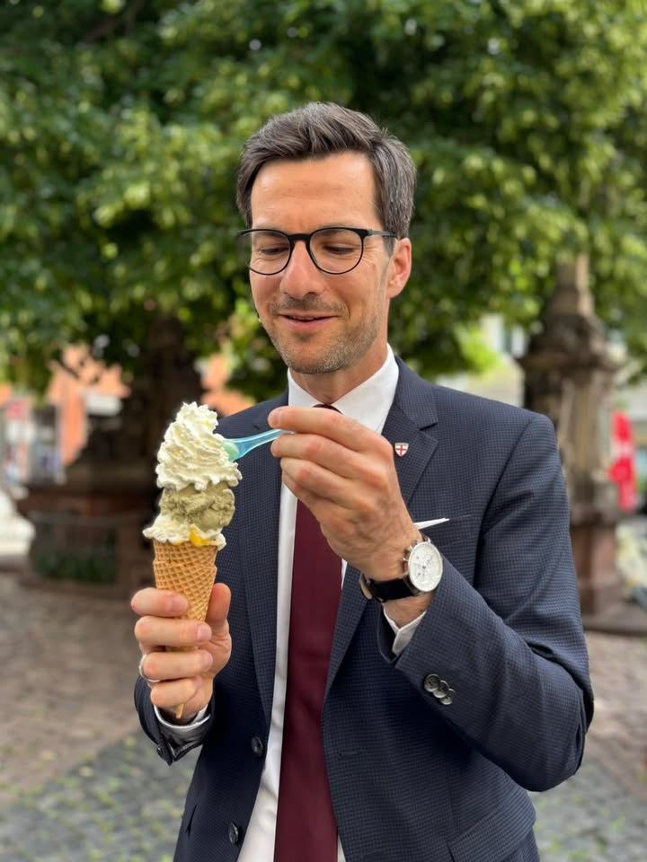 Eine Person in Anzug und Brille hält ein Eis in der Waffel mit zwei Kugeln und Sahne und isst es mit einem blauen Löffel. Im Hintergrund sind Bäume und eine Straßenszene unscharf zu erkennen. (KI-Alternativtext)