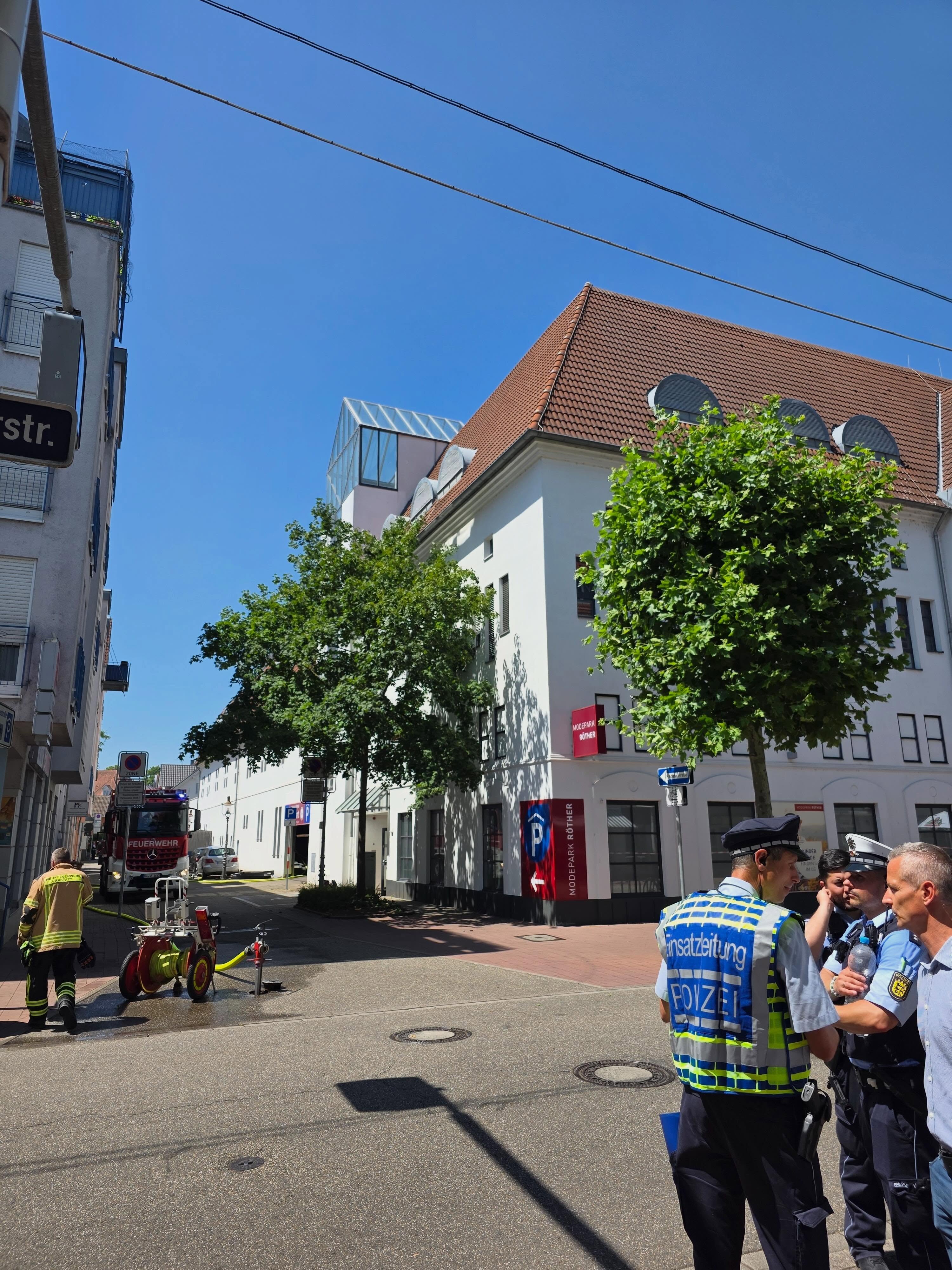 Auf einer sonnigen Straße unterhalten sich rechts mehrere Personen, darunter eine Person in einer Weste mit der Aufschrift "Einsatzleitung POLIZEI" und eine Person in Polizeiuniform. Links geht eine Person in Feuerwehrkleidung auf ein rotes Feuerwehrauto zu, neben dem ein Schlauchwagen steht. Im Hintergrund befindet sich ein weißes Gebäude mit dem Schild "MODEPARK RÖTHER". (KI-Alternativtext)