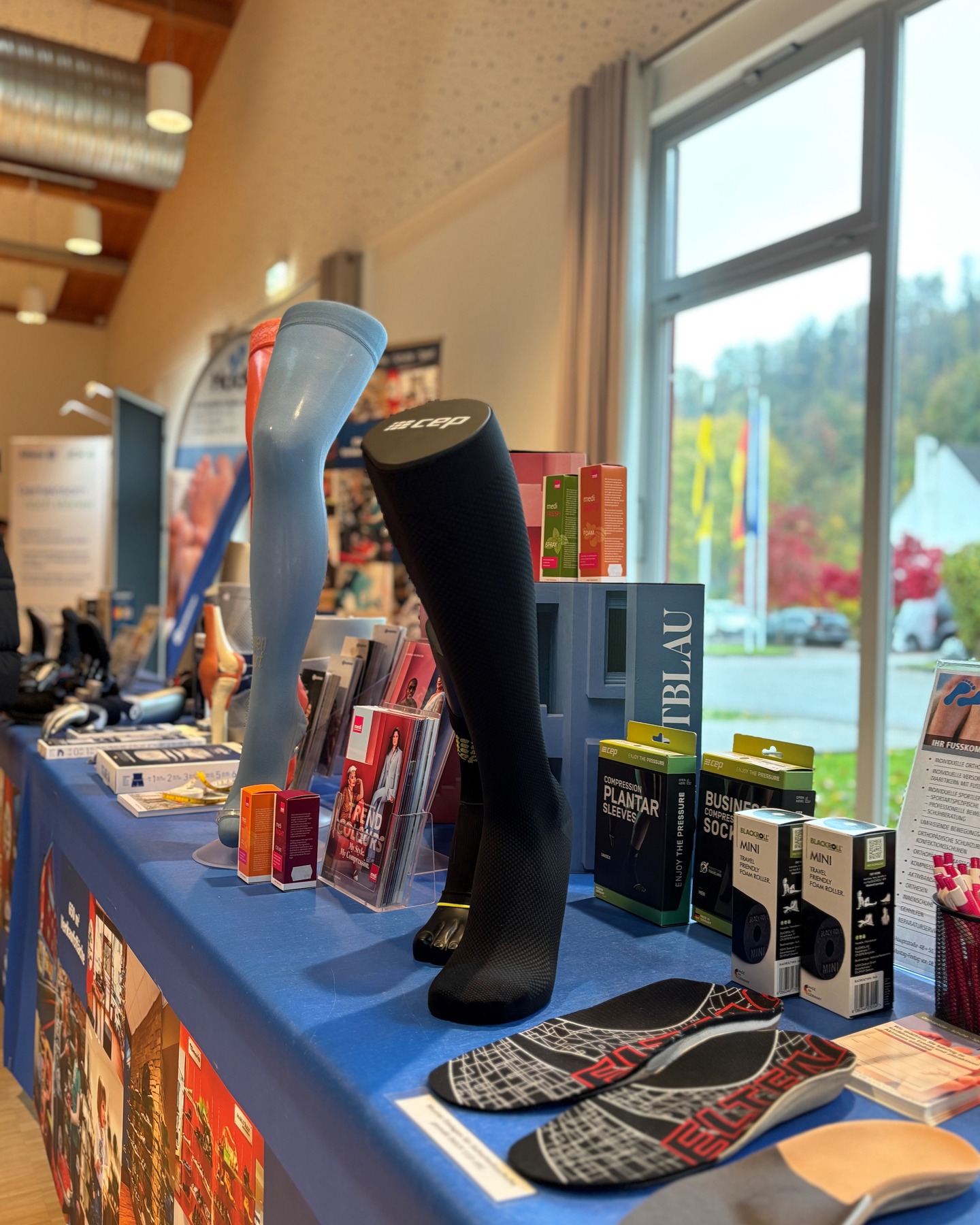 Dieser Alternativtext wurde KI-generiert: Ein Tisch mit verschiedenen Produkten, darunter Kompressionsbekleidung und Fußpflegeartikeln. Im Hintergrund ist durch ein Fenster eine Außenansicht mit Bäumen und einem Parkplatz zu sehen.