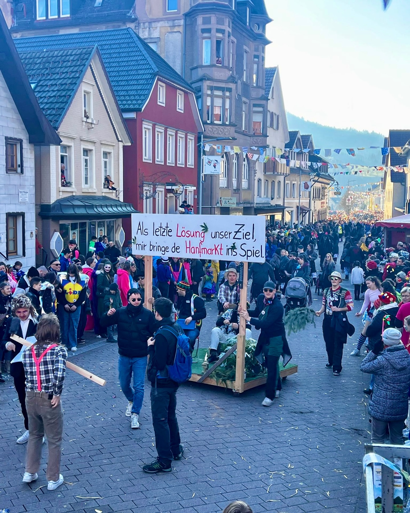 Auf einer Straße in einer Stadt ist eine Parade zu sehen. Ein großes Schild wird bei der Parade mitgetragen.