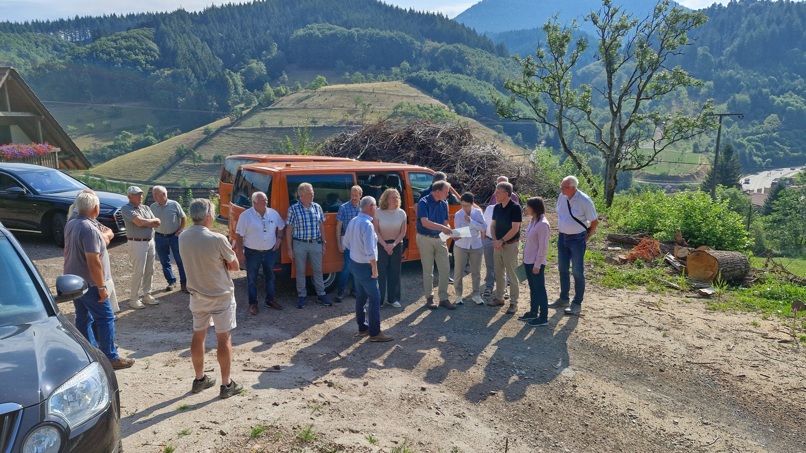 Menschen stehen um einen orangefarbenen Van und hören einer Person zu, die Papiere hält. Links sind weitere Fahrzeuge und ein Haus zu sehen, im Hintergrund erstrecken sich bewaldete Hügel und ein Asthaufen. (KI-Alternativtext)
