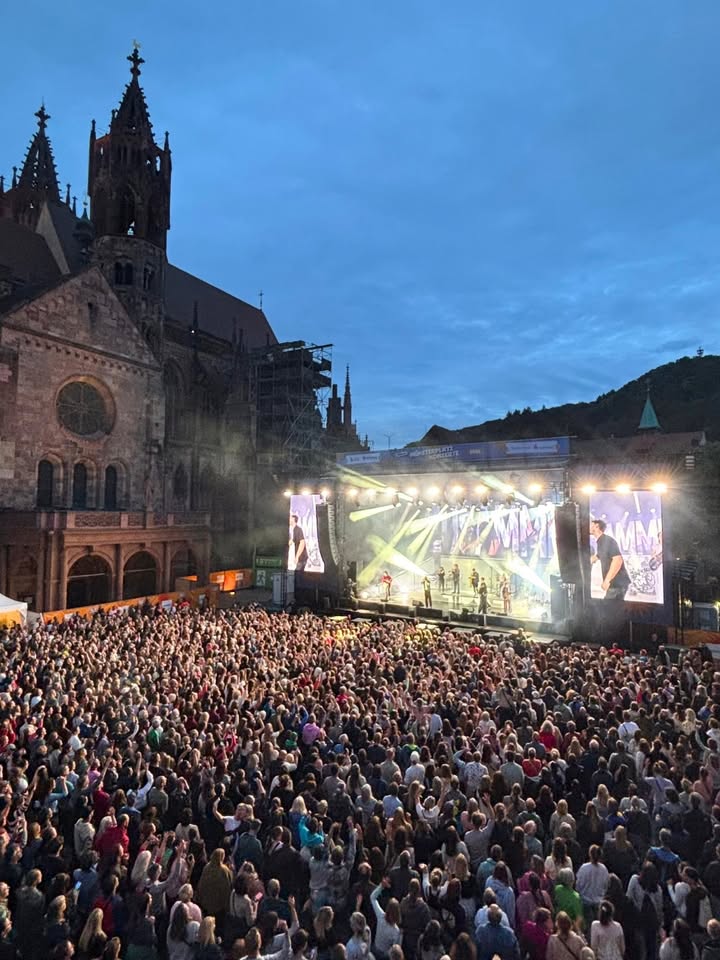 Große Menschenmenge vor einer Bühne mit leuchtenden Scheinwerfern und großen Bildschirmen, im Hintergrund eine gotische Kirche bei Dämmerung. (KI-Alternativtext)
