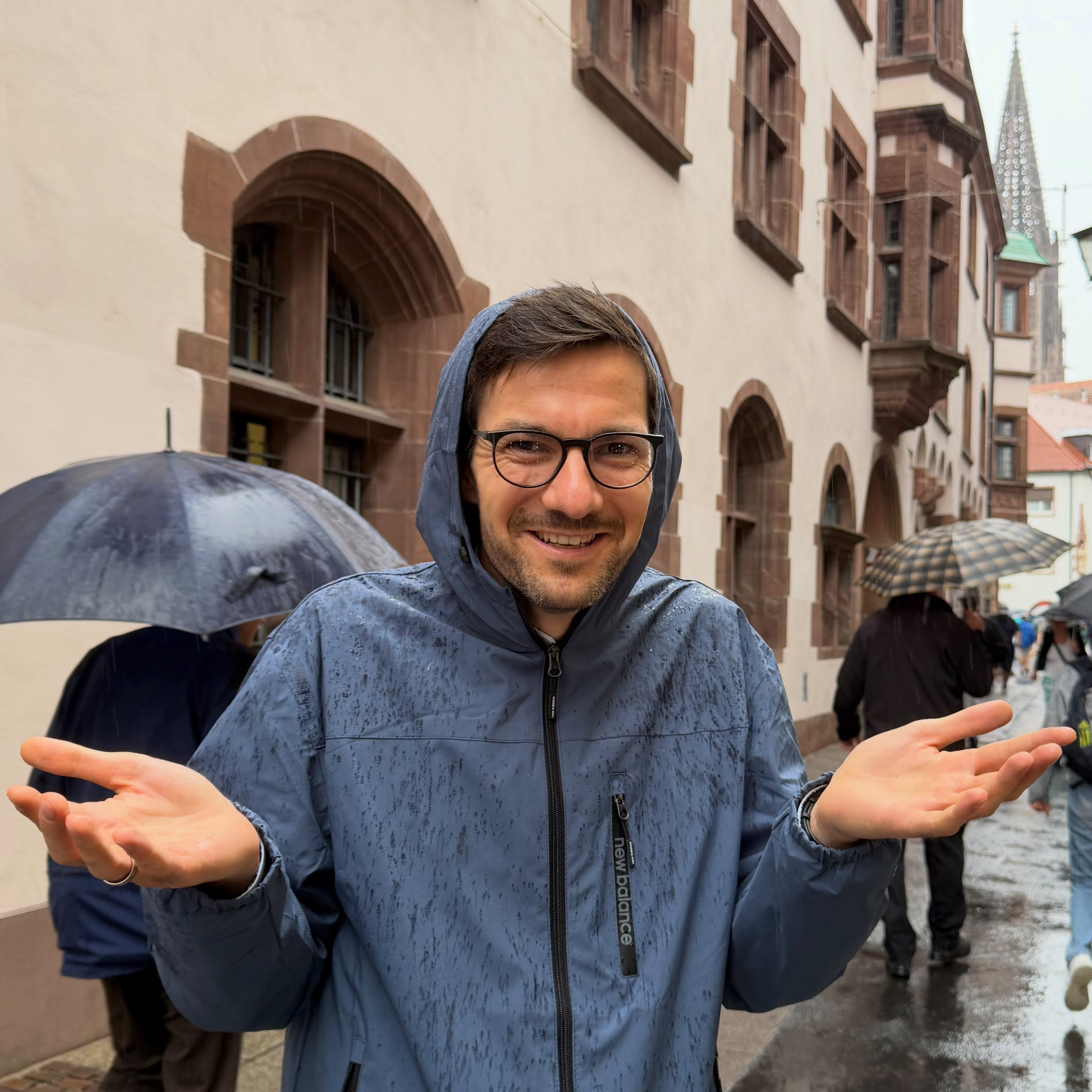 Eine Person in einer blauen, nassen Regenjacke mit Kapuze und Brille steht im Regen und hält beide Hände mit offenen Handflächen nach oben. Auf dem Reißverschluss der Jacke ist der Schriftzug „new balance“ zu sehen. Im Hintergrund sind historische Gebäude und weitere Personen mit Regenschirmen auf einer nassen Straße zu erkennen. (KI-Alternativtext)