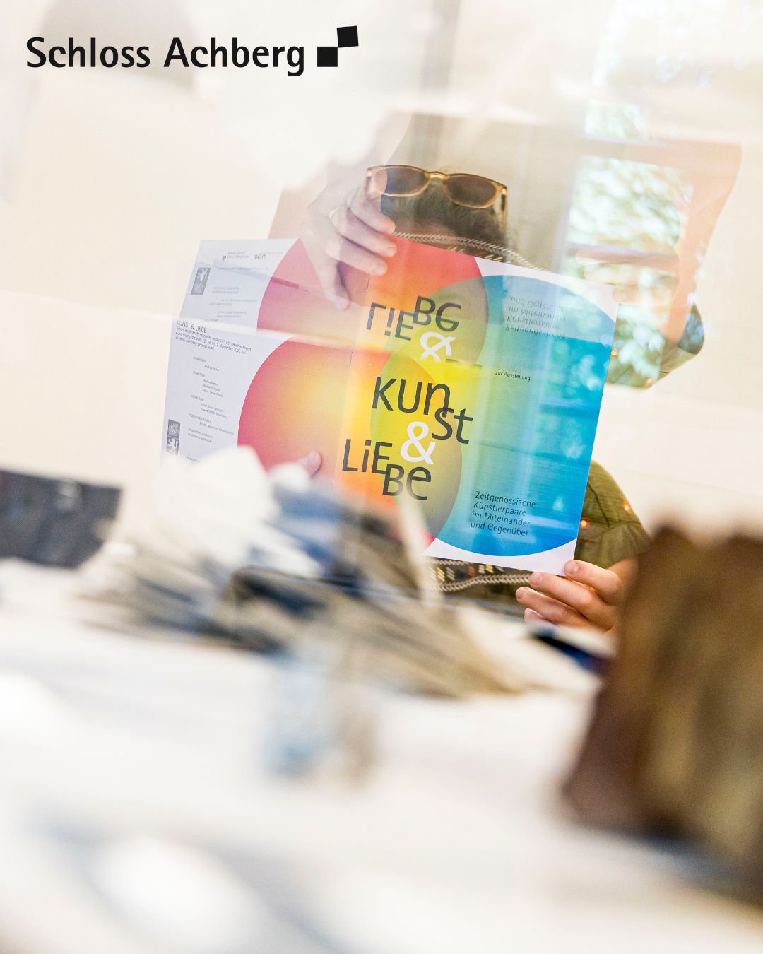 Eine Person mit Sonnenbrille hält eine Broschüre mit dem Titel 'Kunst & Liebe' vor dem Gesicht. Das Bild zeigt das Logo von 'Schloss Achberg'.