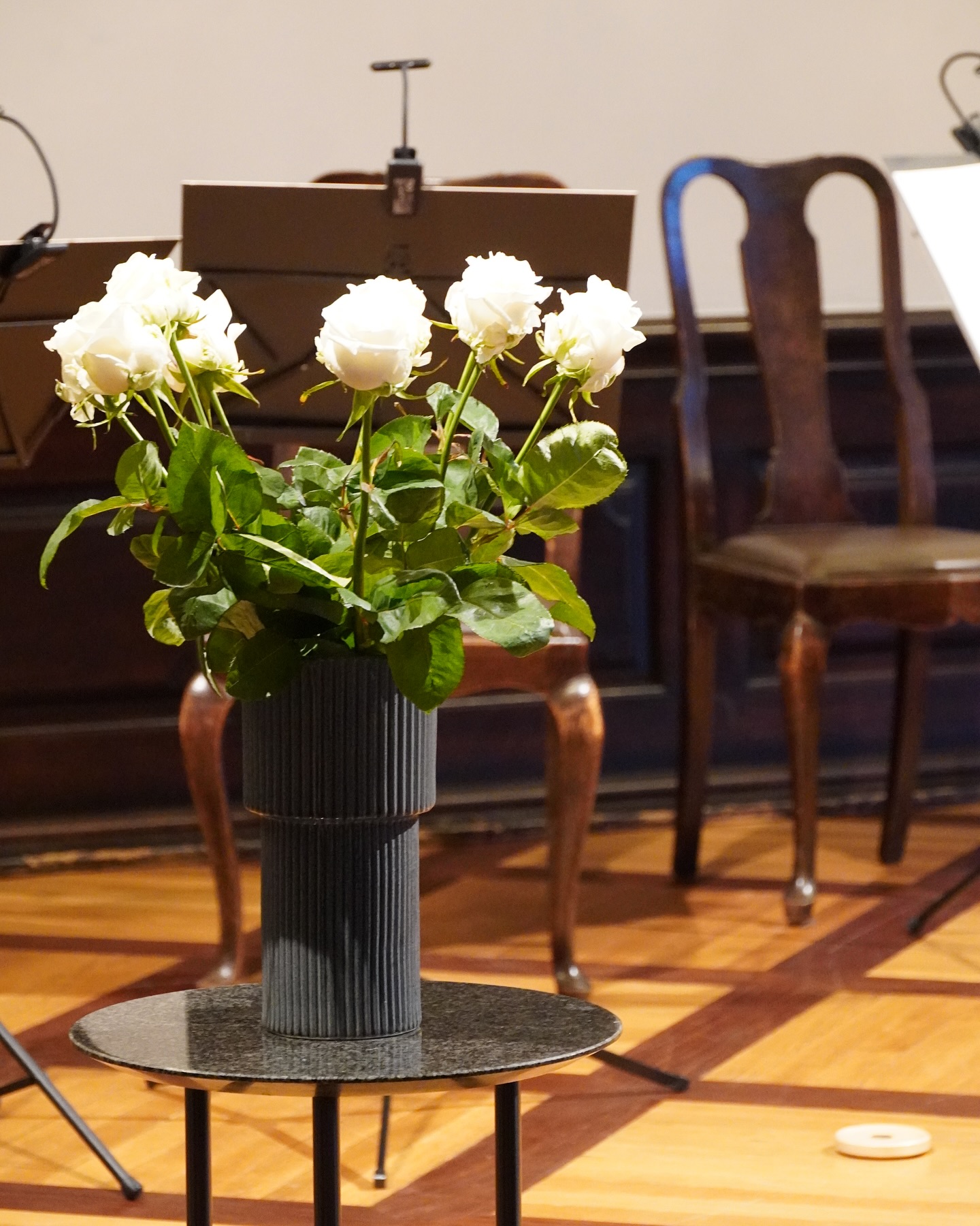 Ein dunkler, gerippter Vasen steht auf einem kleinen Tisch mit einem schwarzen Marmor-Top. Darin befinden sich weiße Rosen.