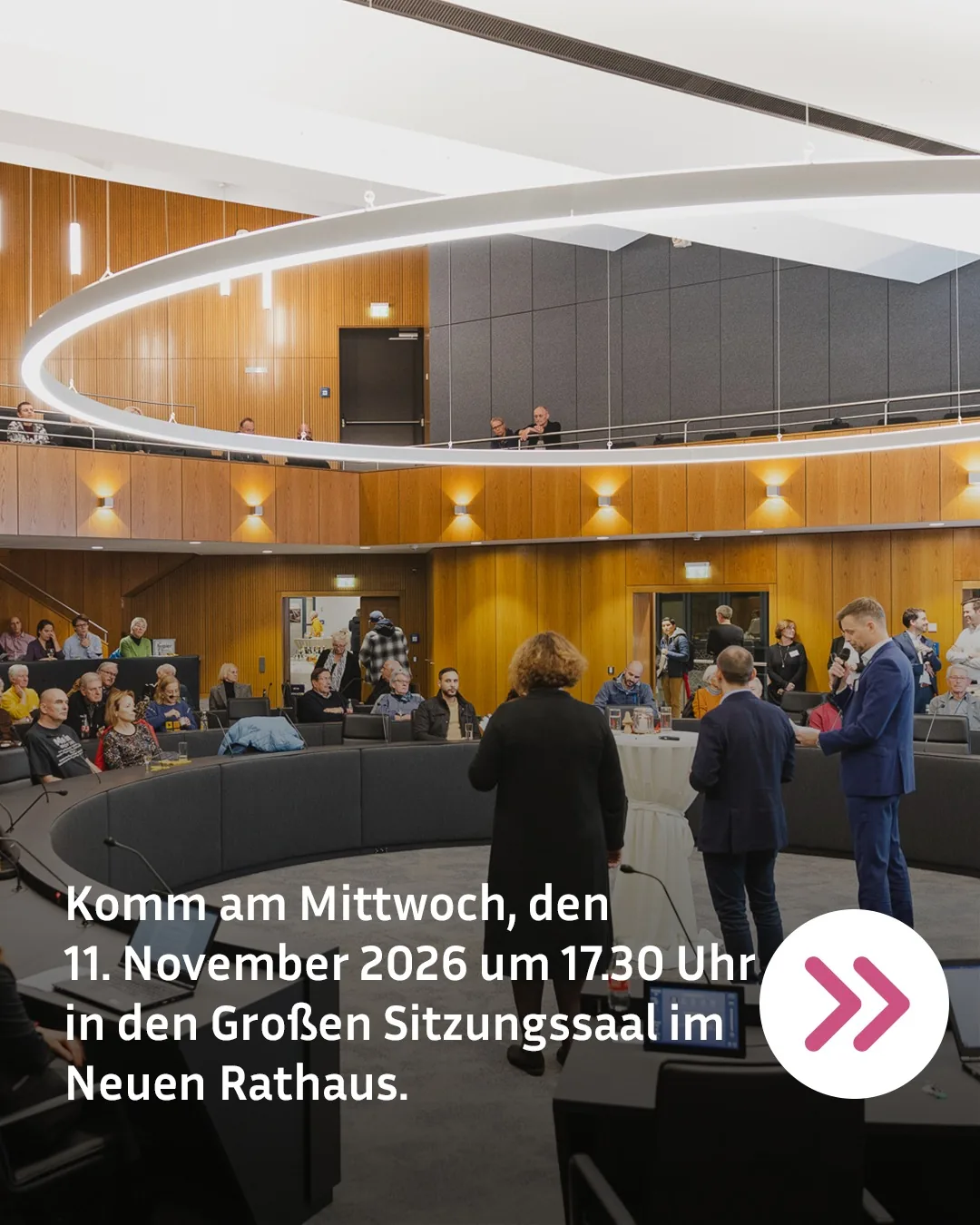 Personen sitzen in einem großen Sitzungssaal mit runder Sitzkonfiguration und einem Balkon im Hintergrund. Drei Personen stehen vor Mikrofonen, eine hält ein Mikrofon in die Hand. Weißer Text am unteren Rand lautet: "Komm am Mittwoch, den 11. November 2026 um 17.30 Uhr in den Großen Sitzungssaal im Neuen Rathaus." Rechts unten befindet sich ein weißer Kreis mit zwei rosa Pfeilen nach rechts. (KI-Alternativtext)
