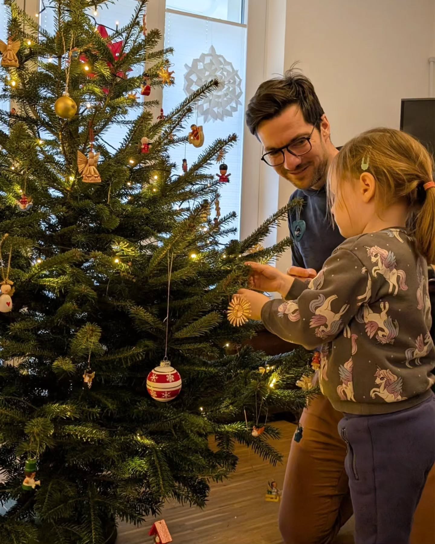 Dieser Alternativtext wurde KI-generiert: Ein geschmückter Weihnachtsbaum steht im Raum. Zwei Personen sind dabei, den Baum mit Ornamenten zu dekorieren.
