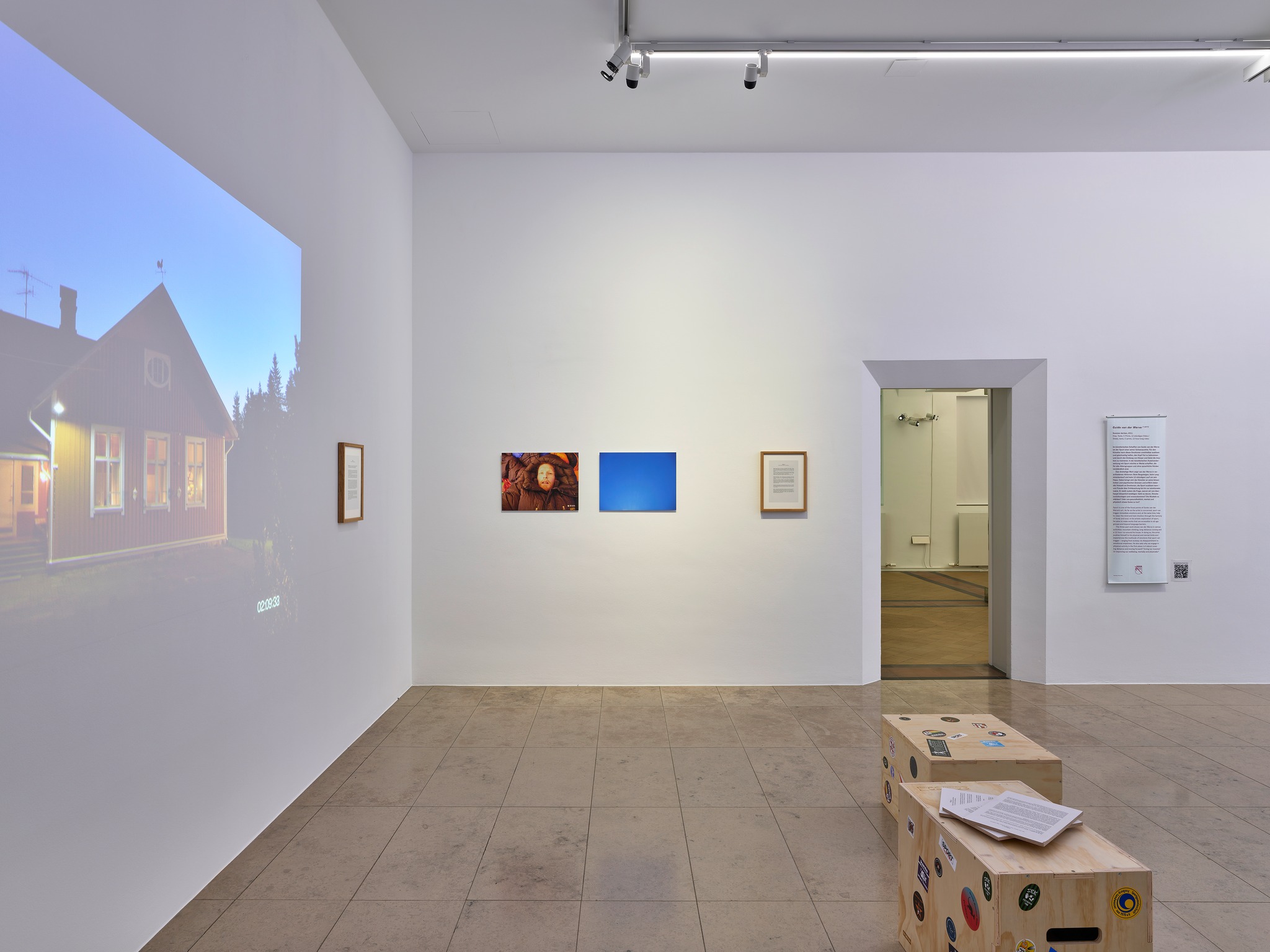 Ein heller Ausstellungsraum mit weißen Wänden, an dem links ein Haus projiziert wird. An der Rückwand hängen drei Kunstwerke: ein Bild einer Person, ein blaues Bild und ein gerahmter Text. Rechts führt eine Tür in einen Nebenraum, daneben hängt ein Textplakat, im Vordergrund stehen zwei Holzkisten mit Aufklebern und Papieren. (KI-Alternativtext)