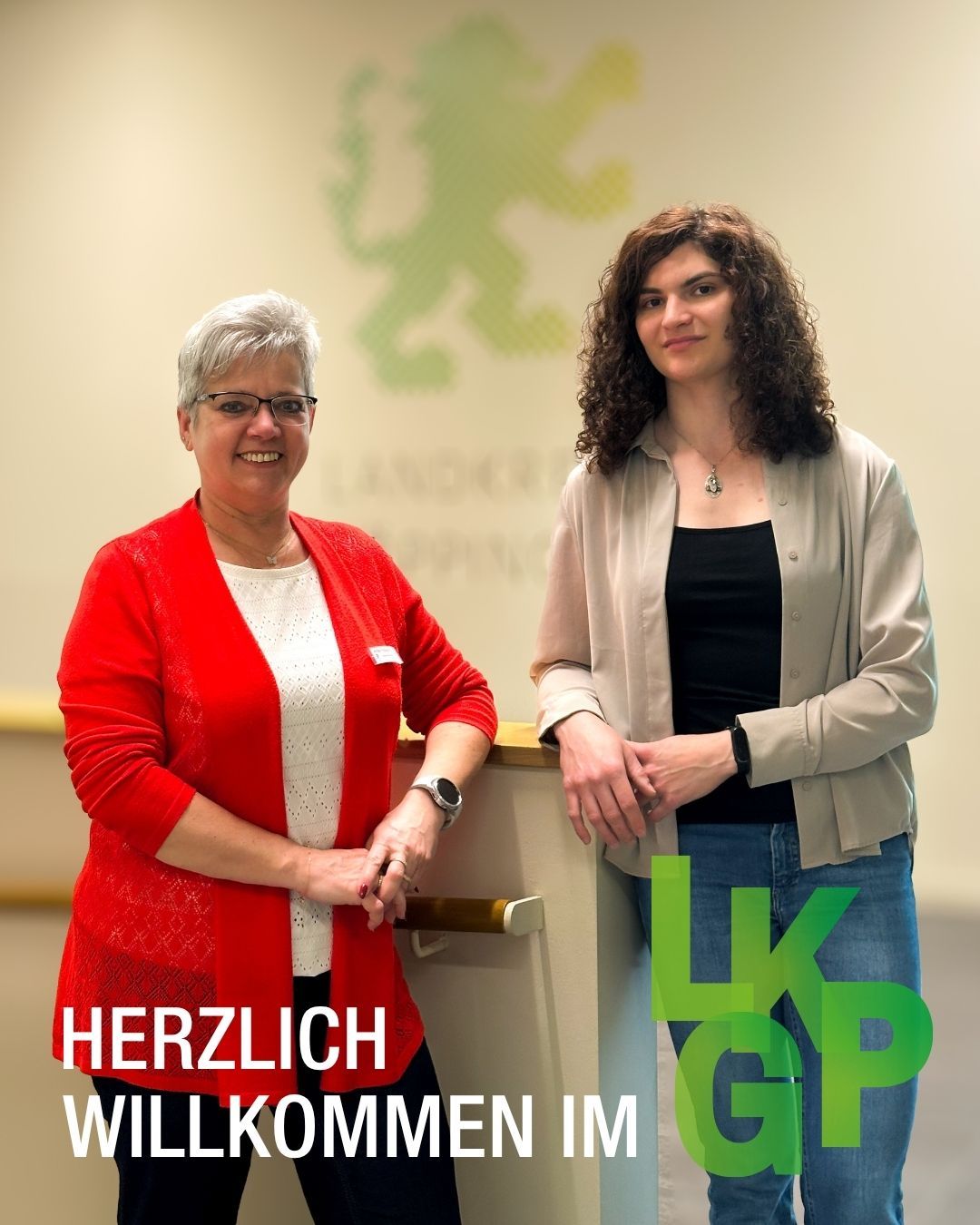 Zwei Personen stehen nebeneinander vor einer Wand mit einem grünen Wappen. Links trägt eine Person eine rote Strickjacke, rechts eine helle Bluse über einem schwarzen Top. Im Vordergrund steht der Text „HERZLICH WILLKOMMEN IM LKGP“. (KI-Alternativtext)