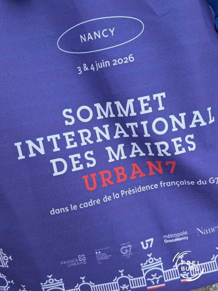 Blauer Stoff mit weißem und rotem Text: „NANCY, 3 & 4 juin 2026, SOMMET INTERNATIONAL DES MAIRES, URBAN7, dans le cadre de la Présidence française du G7“. Unten Logos von France Urbaine, G7 France, U7, métropole GrandNancy und Freiburg. (KI-Alternativtext)