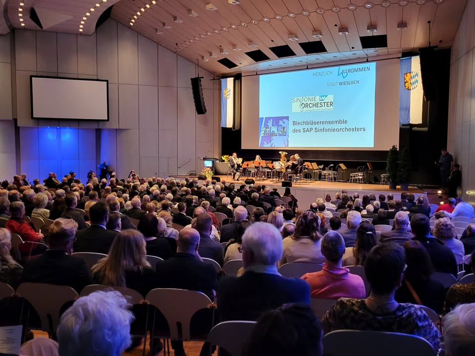Dieser Alternativtext wurde KI-generiert: Ein Publikum sitzt in einem großen Saal vor einer Bühne. Auf einer Leinwand im Hintergrund ist ein Text und ein Bild zu sehen.