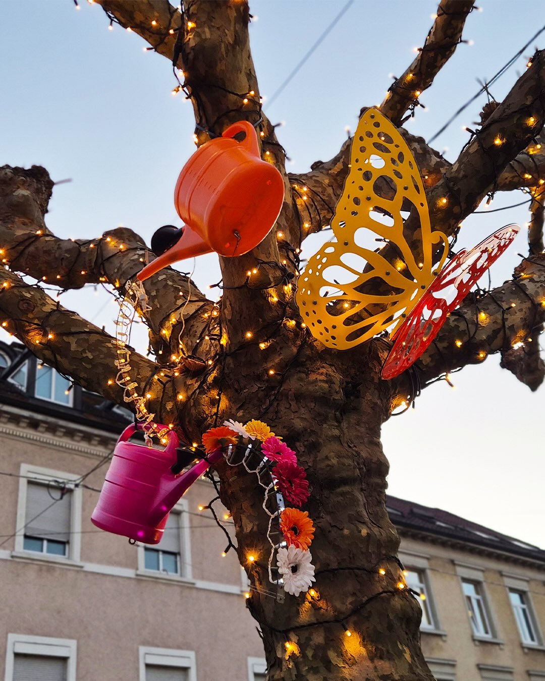 Dieser Alternativtext wurde KI-generiert: Ein Baum ist mit Lichtern, bunten Dekorationen wie Schmetterlingen und Gießkannen geschmückt. Die Aufnahme ist farbig und zeigt eine stimmungsvolle Szene.