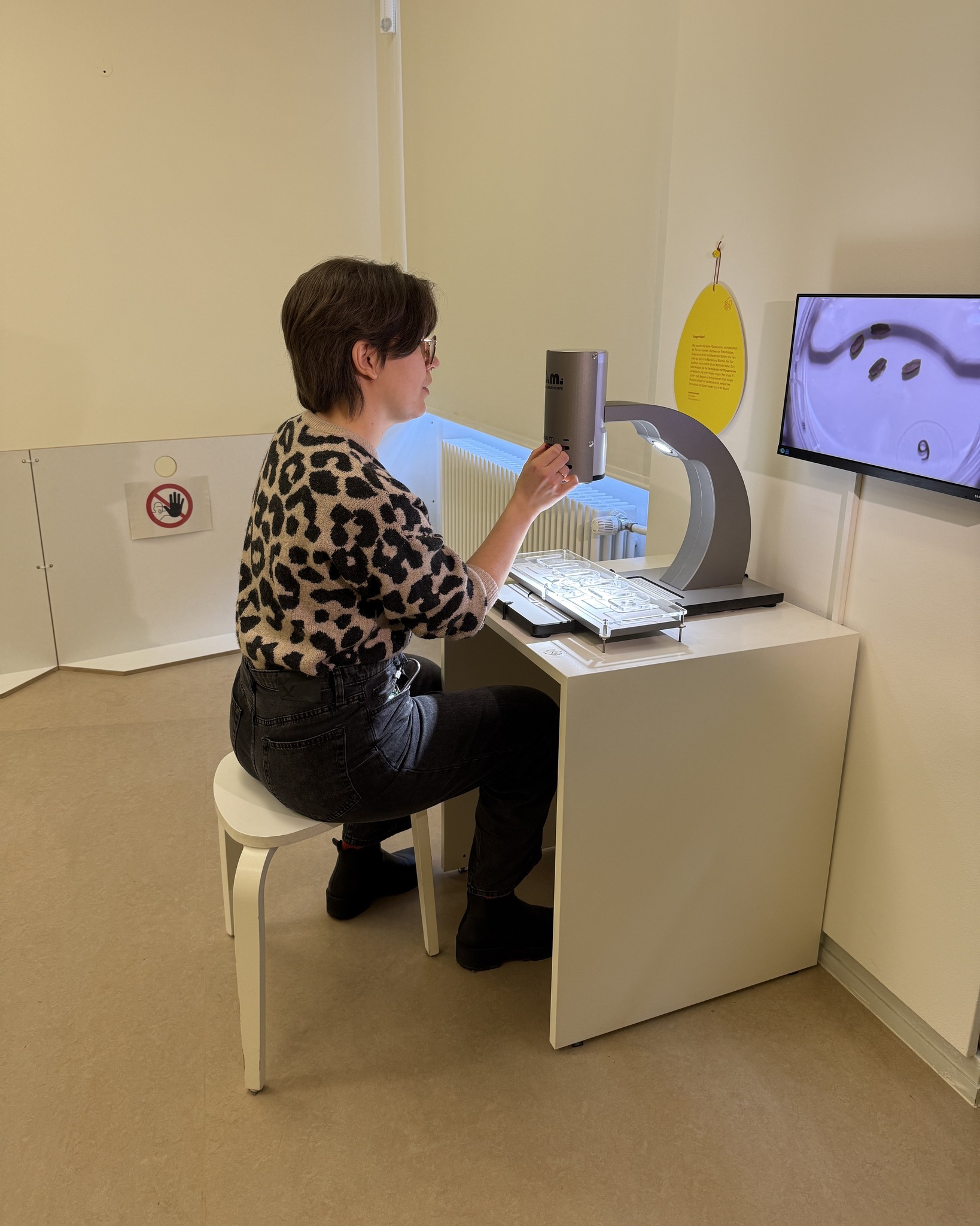 Dieser Alternativtext wurde KI-generiert: Eine Person sitzt vor einem Gerät mit einem Bildschirm und betrachtet etwas auf einer Arbeitsfläche. Im Hintergrund befindet sich eine Tür mit einem Warnhinweis-Schild.