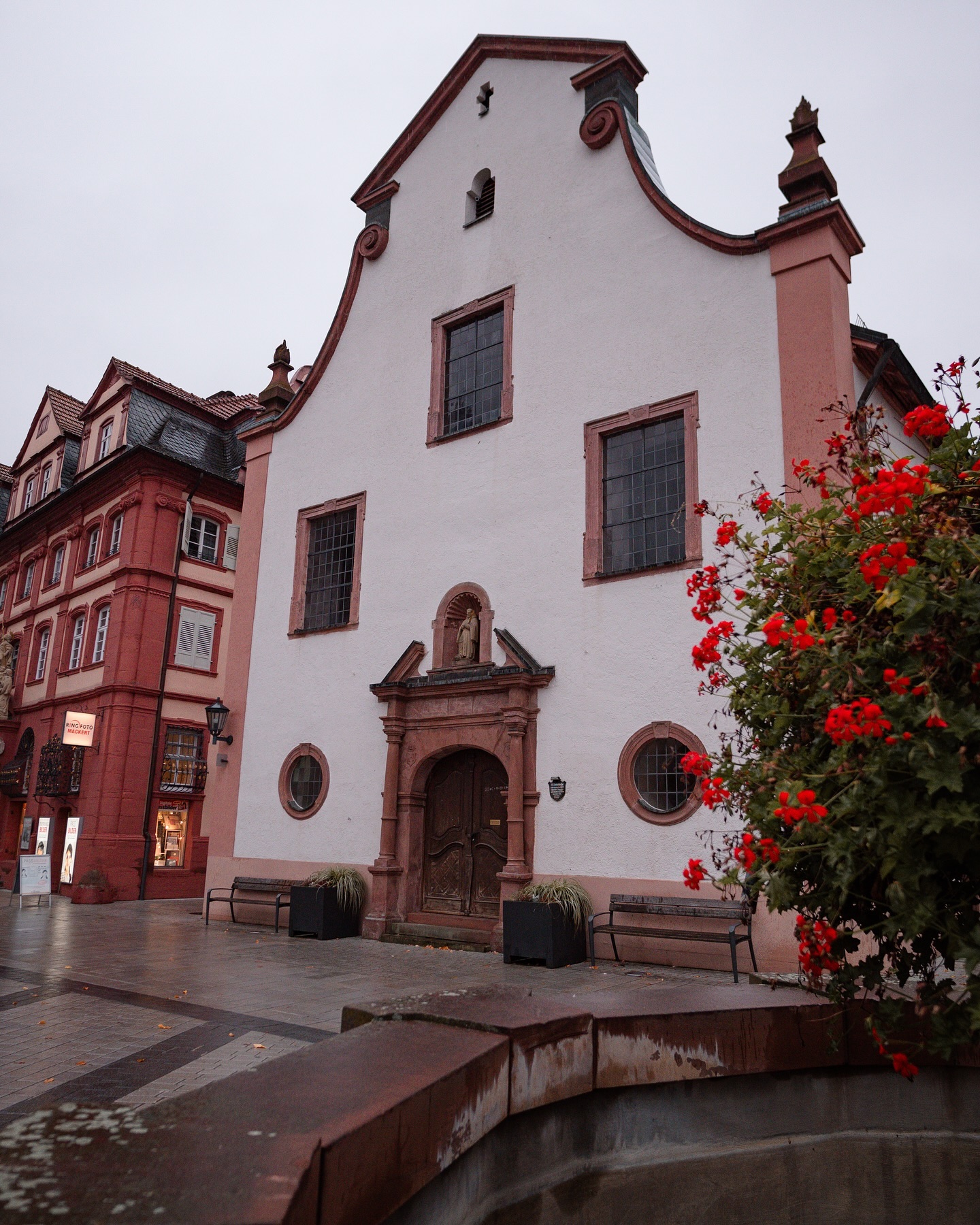 Ein weißes Gebäude mit roten Akzenten und mehreren Fenstern steht neben einem roten Gebäude. Vor dem weißen Gebäude befinden sich Bänke und ein blühender Strauch.