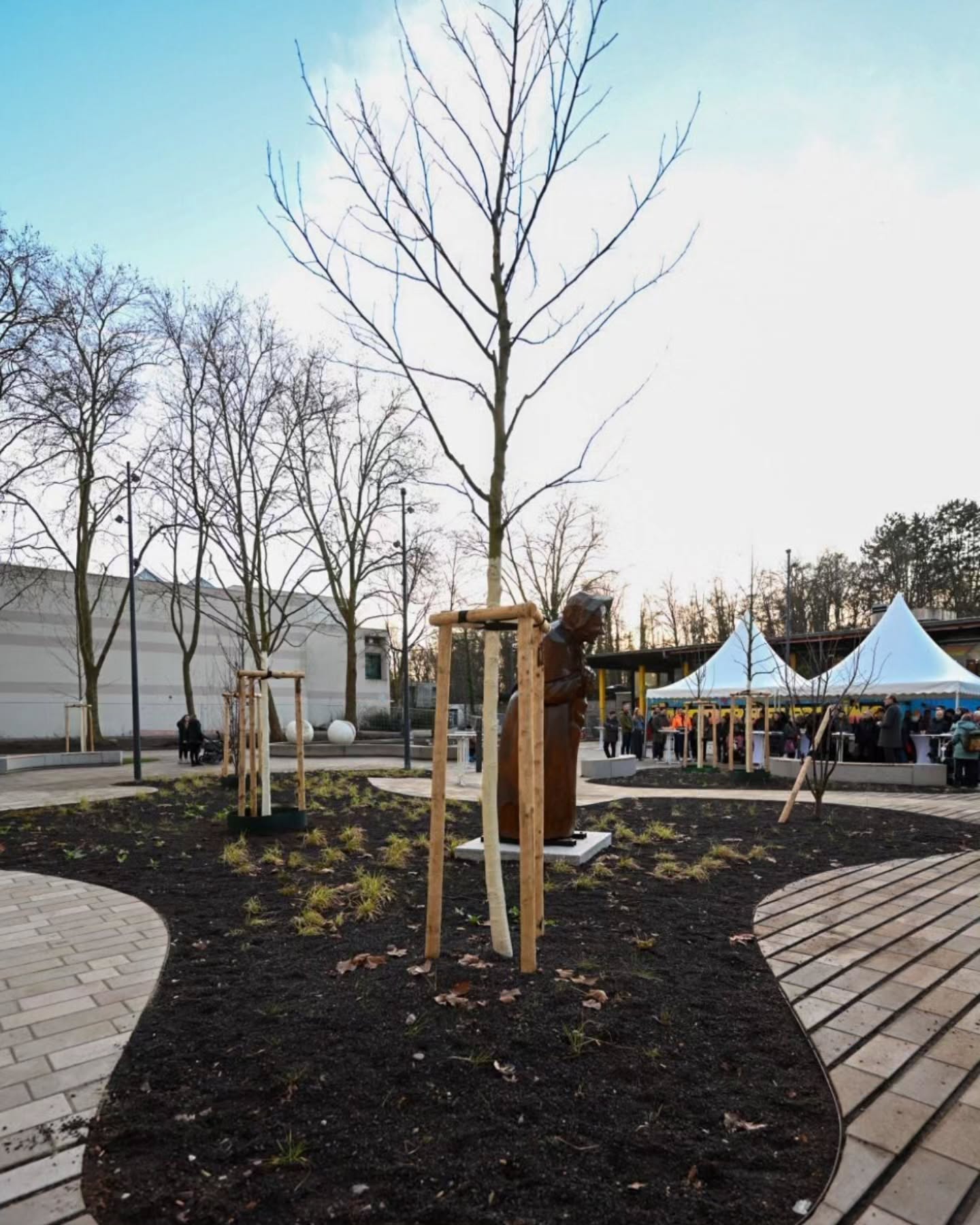 Dieser Alternativtext wurde KI-generiert: Ein weitläufiger Platz mit Bäumen, einer Skulptur und mehreren Zelten im Hintergrund. Der Bereich ist mit Pflastersteinen und frischem Mulch gestaltet.