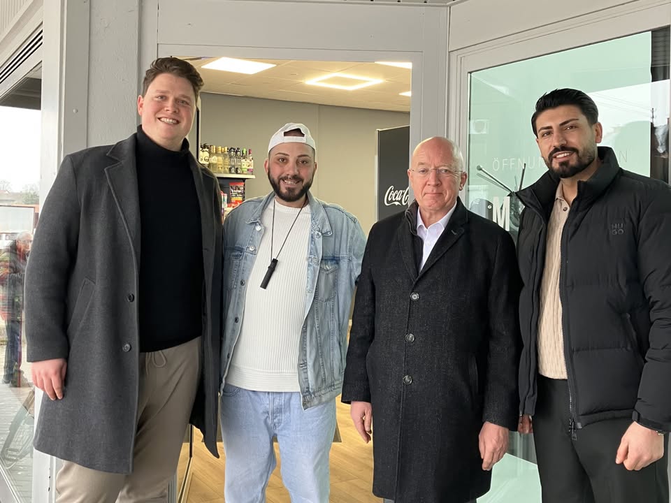 Dieser Alternativtext wurde KI-generiert: Eine Gruppe von vier Personen steht vor einer gläsernen Tür und einem Schaufenster. Im Hintergrund sind ein Raum und ein Getränkeautomat zu sehen.