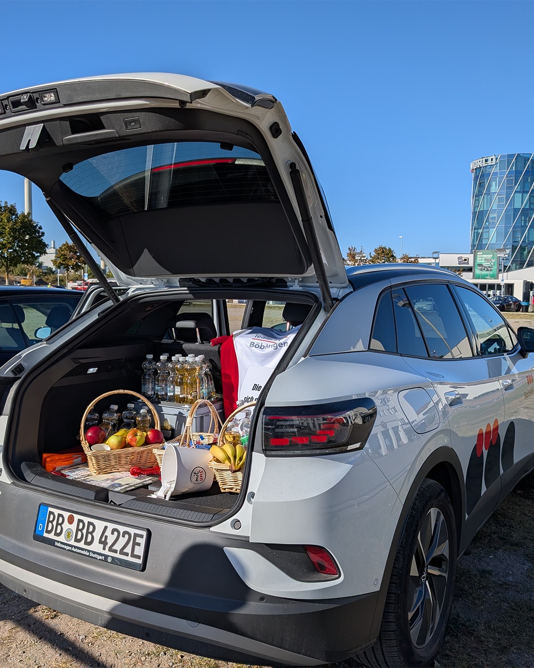 Ein weißes Auto mit geöffnetem Kofferraum steht im Freien unter blauem Himmel. Im Kofferraum stehen Obstkörbe, Flaschen, Getränkeboxen und Tische bereit. Das Kennzeichen zeigt "BB BB 422E" sowie den Schriftzug "Volkswagen Automobile Stuttgart". (KI-Alternativtext)
