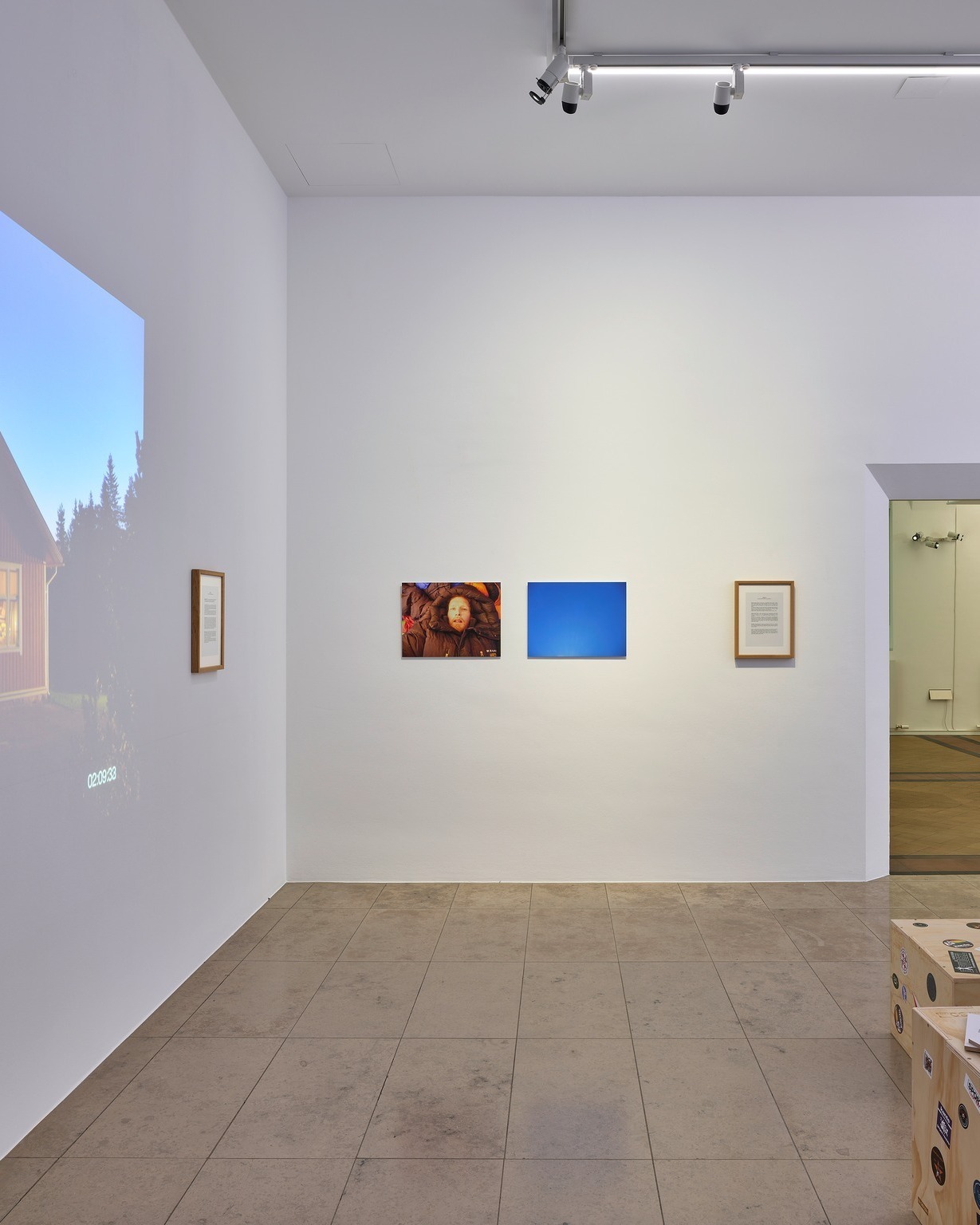 Ein heller Ausstellungsraum mit weißen Wänden und Fliesenboden. An der linken Wand ist eine Projektion eines Hauses und Bäume mit der Zeitangabe 02:03:33 sowie ein gerahmter Text zu sehen. An der Rückwand hängen drei Kunstwerke nebeneinander: ein Foto einer Person im Wintermantel, ein reines blaues Rechteck und ein weiterer gerahmter Text. Rechts unten stehen Holzkisten. (KI-Alternativtext)