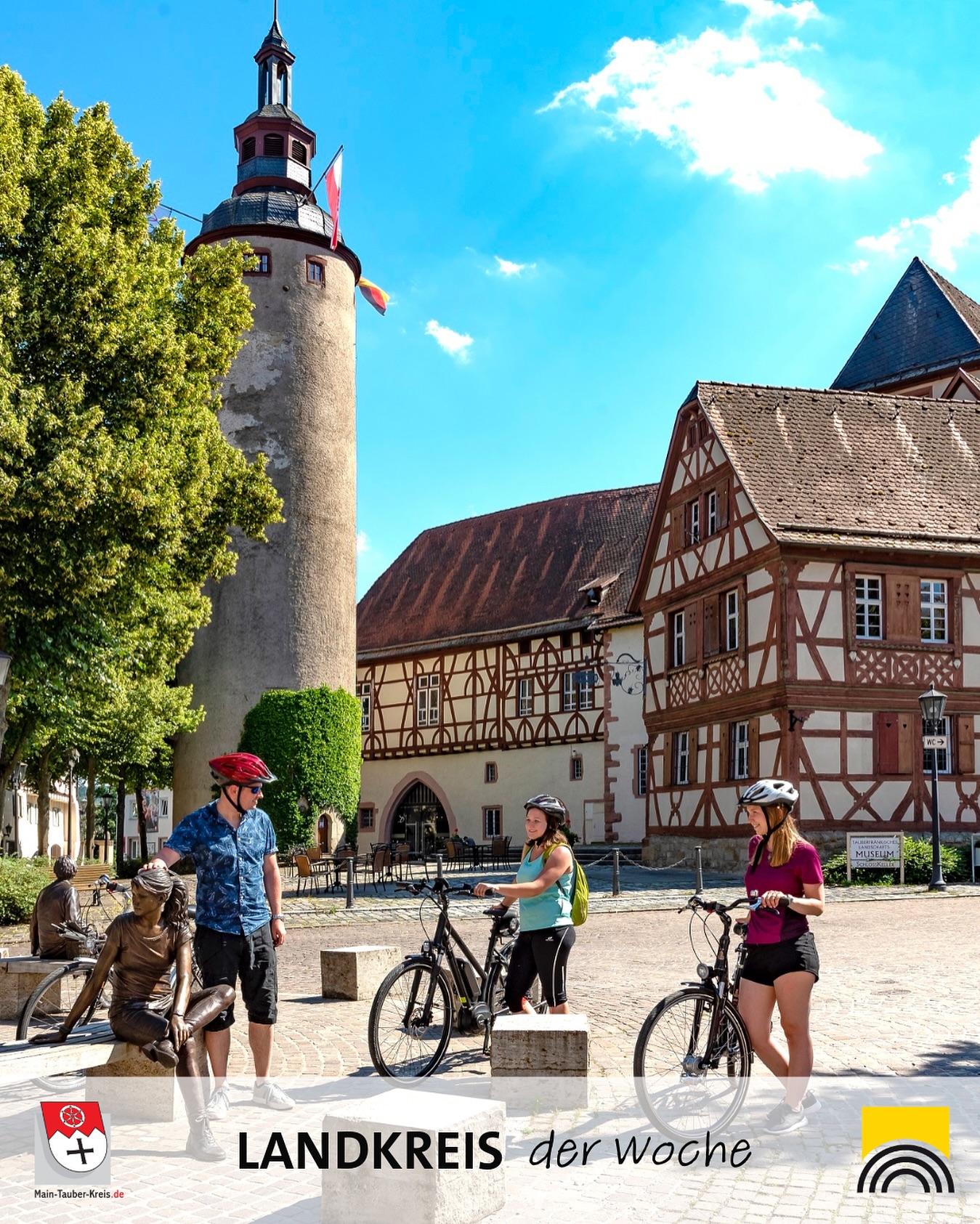 Fahrradfahrer*innen an einem historischen Platz mit Fachwerkhäusern und einem Turm. Sonniger Tag.
