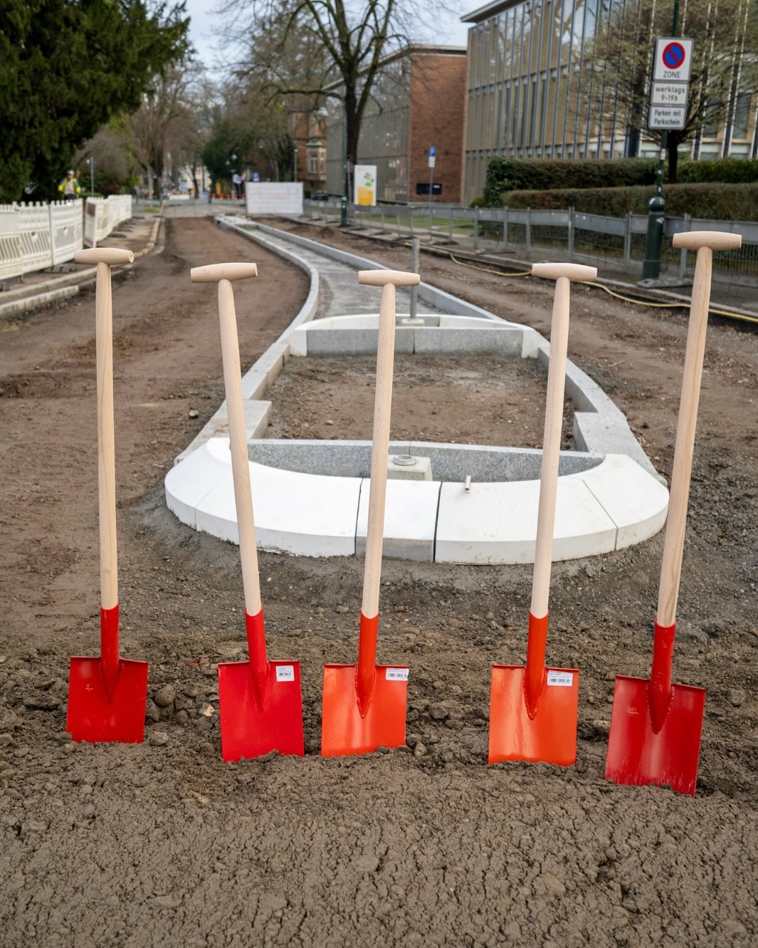 Dieser Alternativtext wurde KI-generiert: Ein Straßenabschnitt befindet sich im Umbau, umgeben von Schaufeln. Im Hintergrund sind Gebäude und Verkehrsschilder zu sehen.