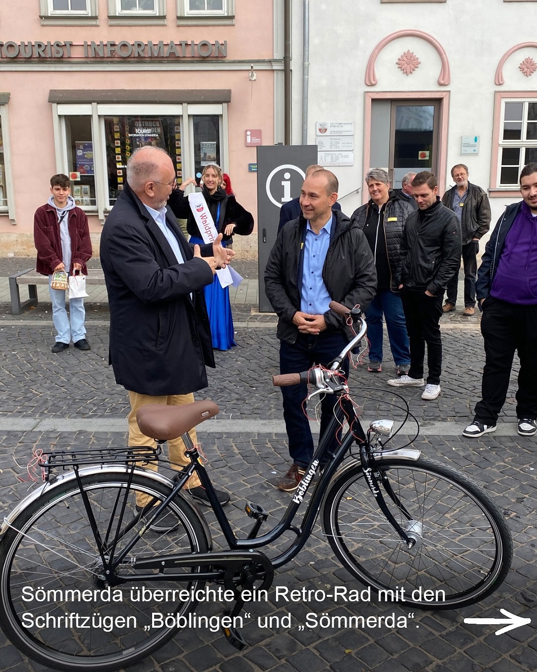 Dieser Alternativtext wurde KI-generiert: Schwarz-weißes Foto einer Straßenszene. Mehrere Personen stehen und sitzen vor einer Fassade mit Schaufenstern und einem Fahrrad, wobei ein Dokument überreicht wird.