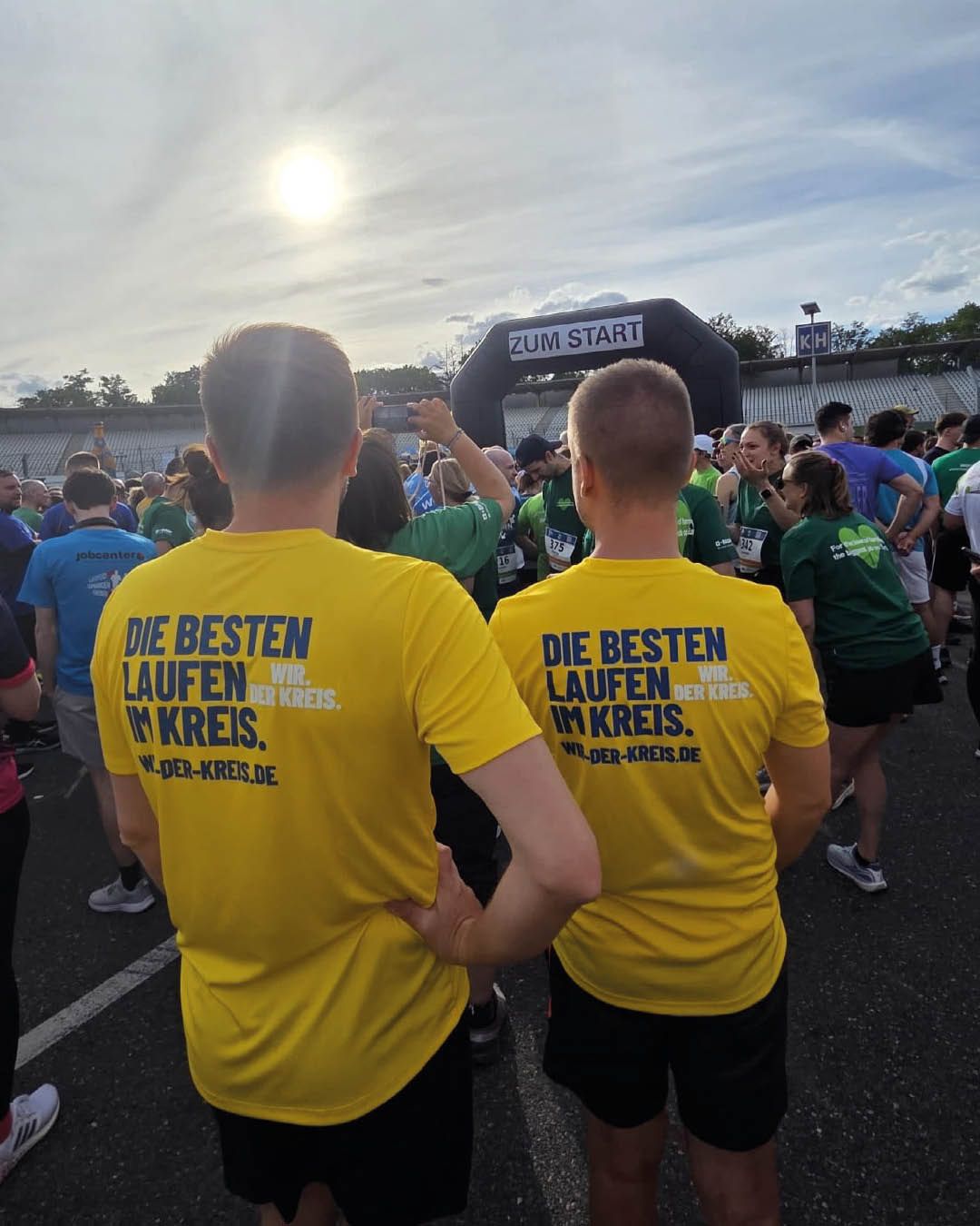Zwei Personen in gelben T-Shirts mit dem Aufdruck „DIE BESTEN LAUFEN WIR. IM KREIS. WIR-DER-KREIS.DE“ stehen vor einem Startbogen mit der Aufschrift „ZUM START“. Im Hintergrund warten weitere Läufer auf den Start, die Sonne steht tief am Himmel. (KI-Alternativtext)