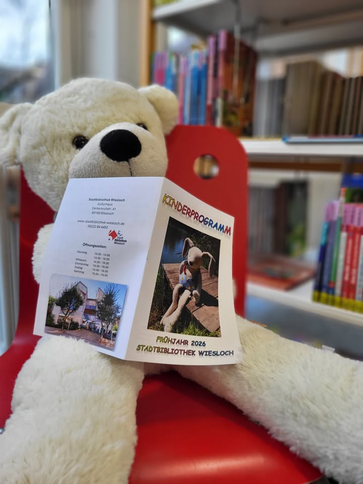 Dieser Alternativtext wurde KI-generiert: Ein Stofftier sitzt vor einem Bücherregal und einem Werbeplakat. Im Hintergrund ist ein Schild mit Informationen zu einem Kinderprogramm zu sehen.